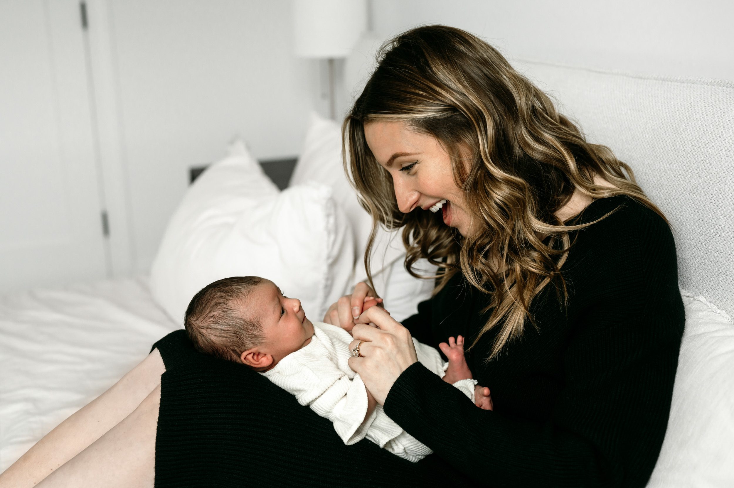 a new mother sitting on her bed with her newborn baby girl on her legs and smiling down at her during an in home lifestyle newborn session