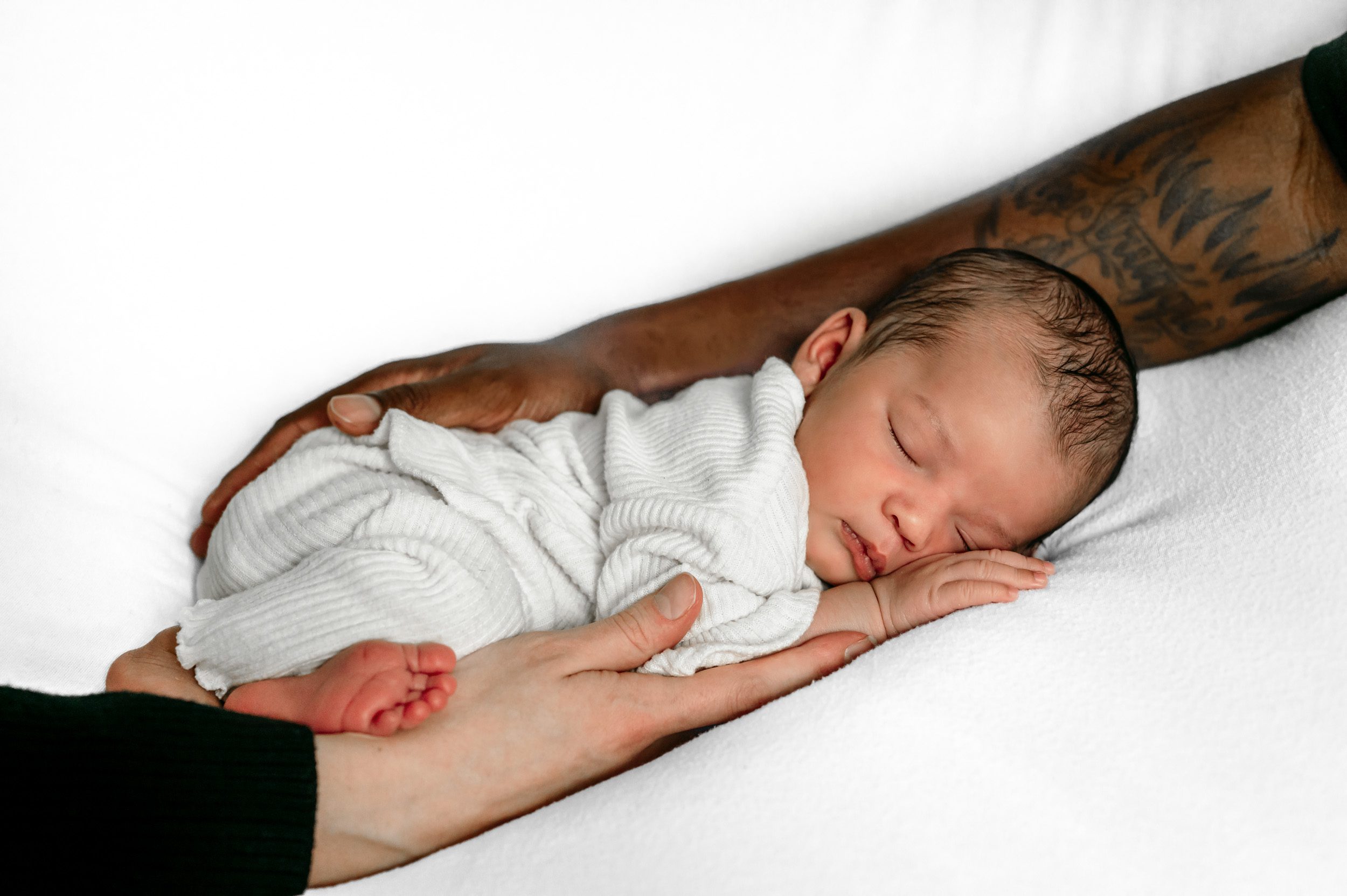 a newborn baby girl laying on her stomach on a white backdrop with her mom and dad's arms cradling her during a newborn photoshoot