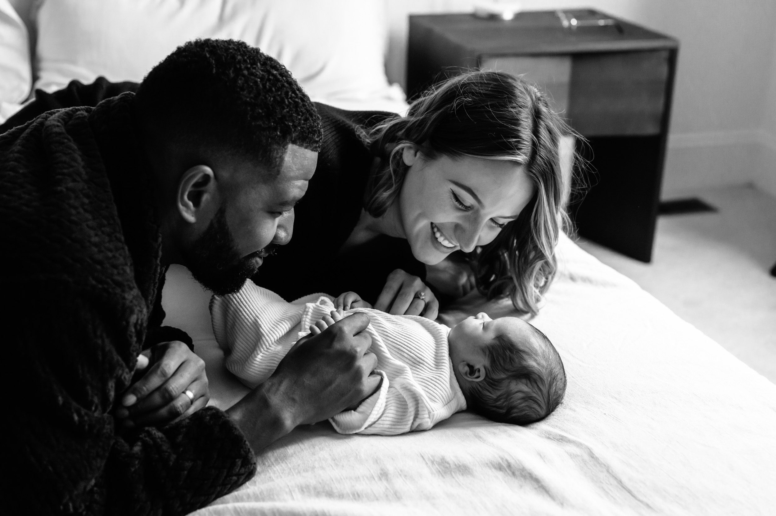 a black and white image of new parents laying on their bed and smiling down at their baby girl during a Phoenixville in home lifestyle newborn photoshoot