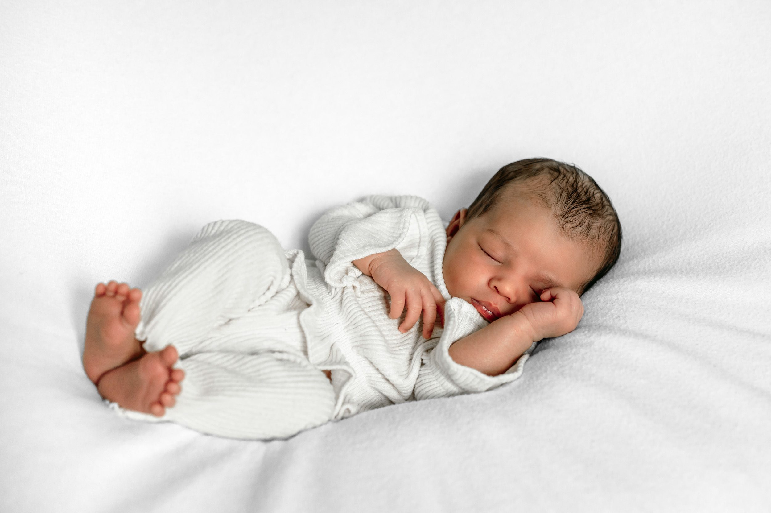 a newborn baby girl laying on her back on a white backdrop with her hands tucked up next to her face during a newborn photoshoot