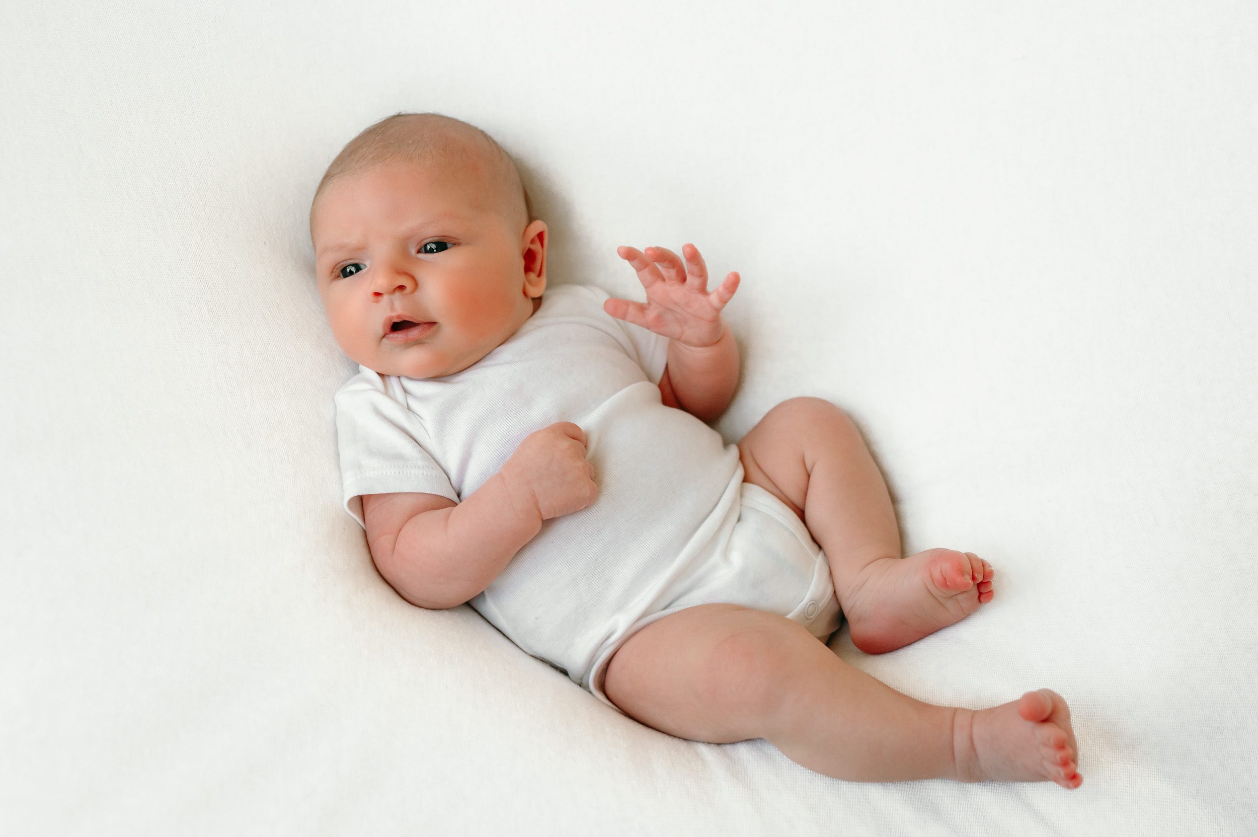  a newborn baby boy in a white onesie on a white backdrop looking toward the window light during a home newborn photoshoot