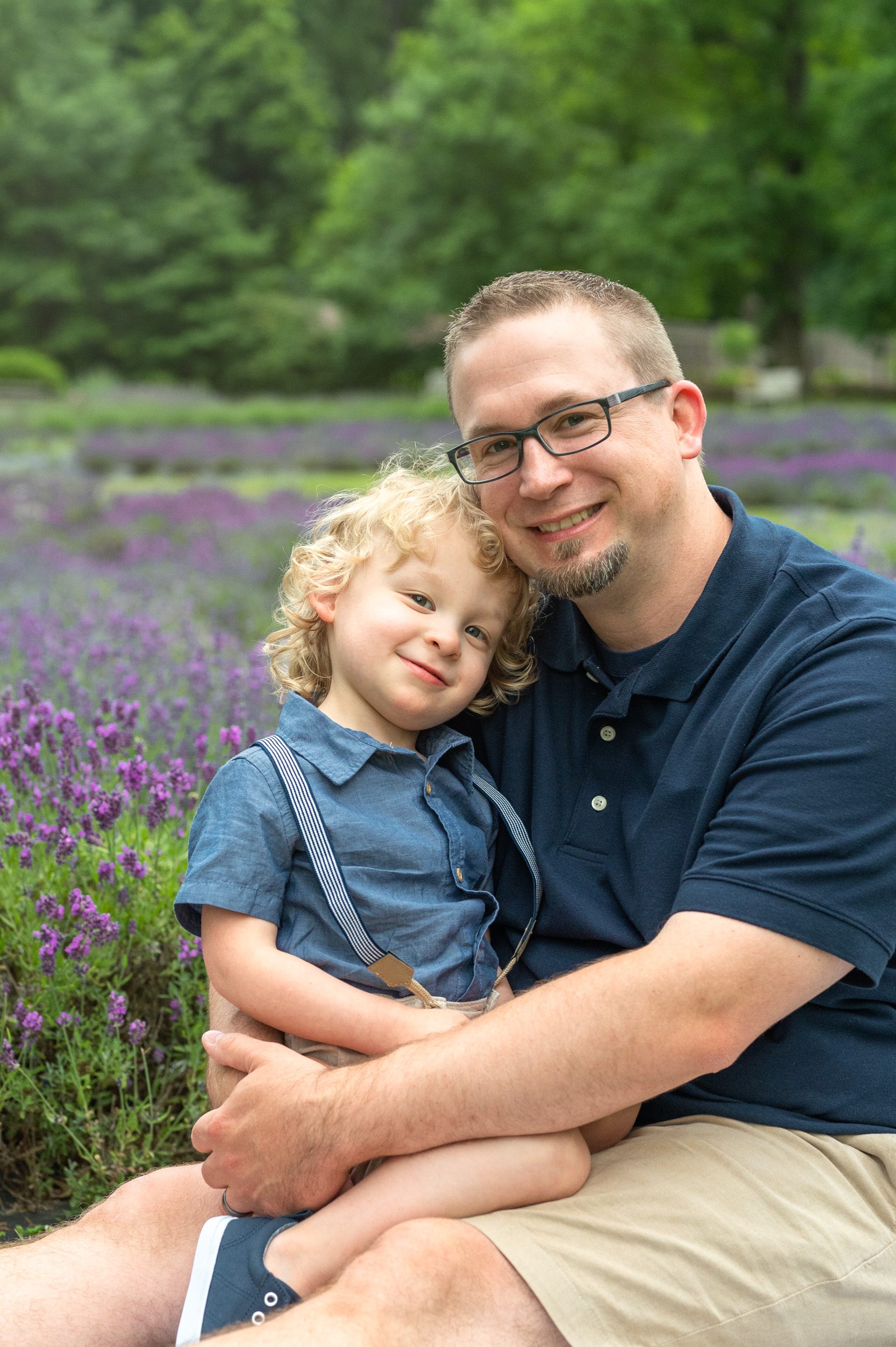 a little boy sitting on his dad's lap and smiling at the camera in a field of purple lavender 