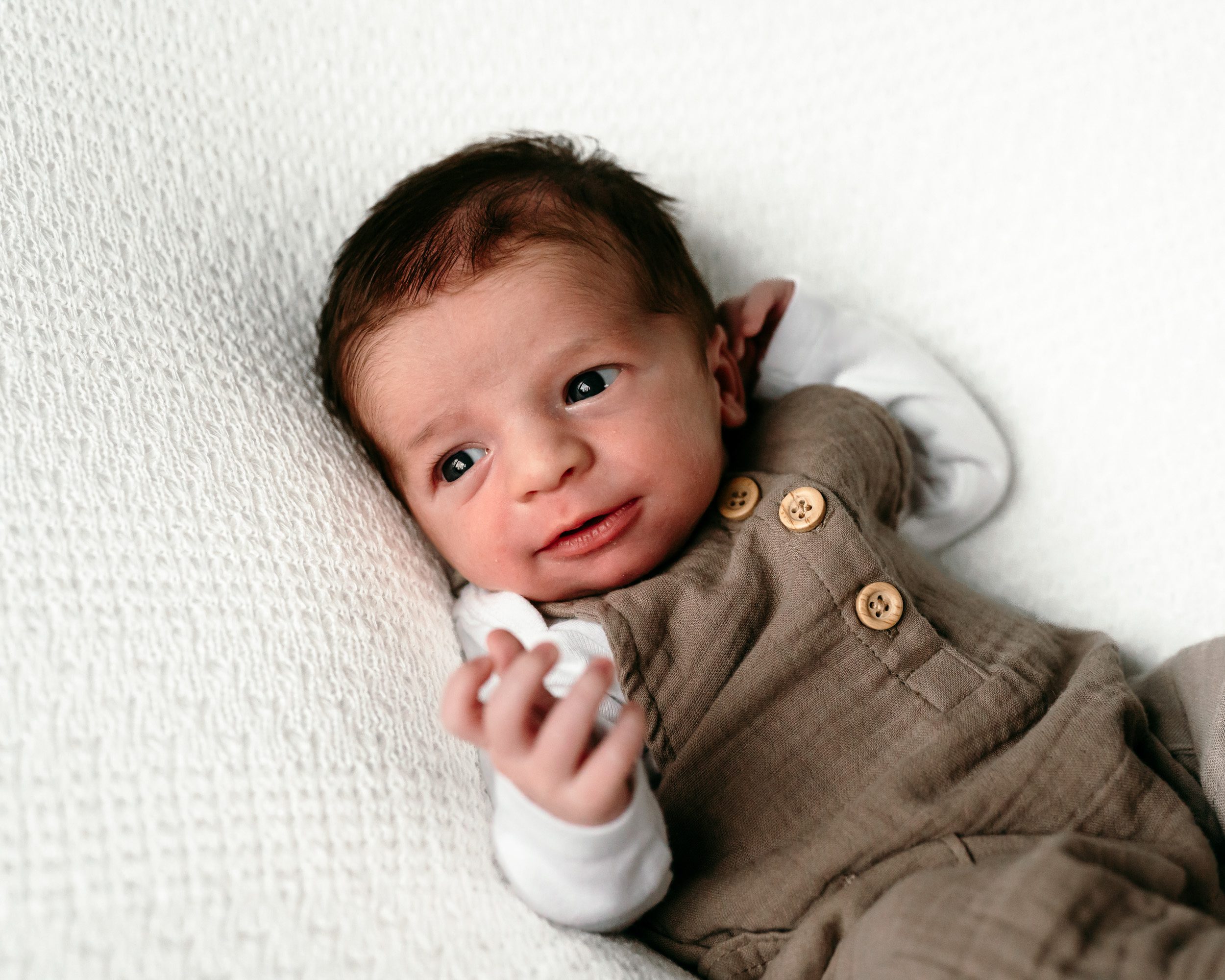 A newborn baby boy laying on a white backdrop and looking off toward the window during a newborn photo session
