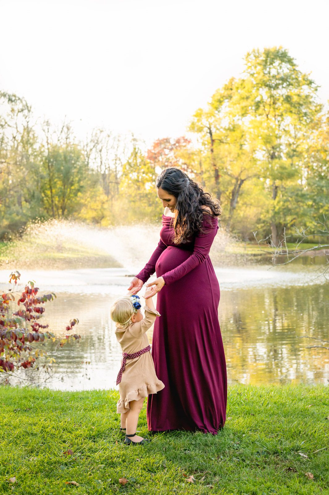 an expecting mom holding hands with her young daughter as they both smile at her belly during a maternity photo session