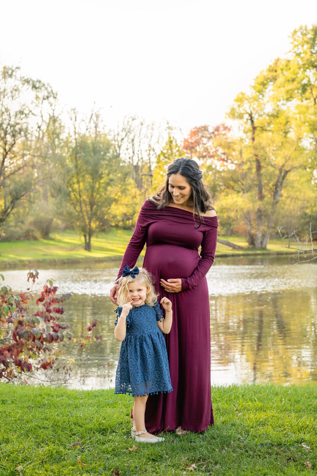 an expecting mom cradling her belly and smiling down at her young daughter while she makes a silly face at the camera during a maternity photoshoot