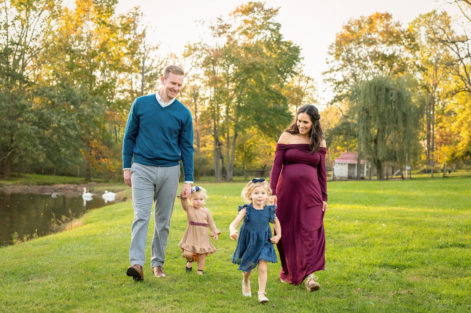 An expecting couple and their two young daughters walking in the grass next to a pond during a phoenixville maternity photoshoot
