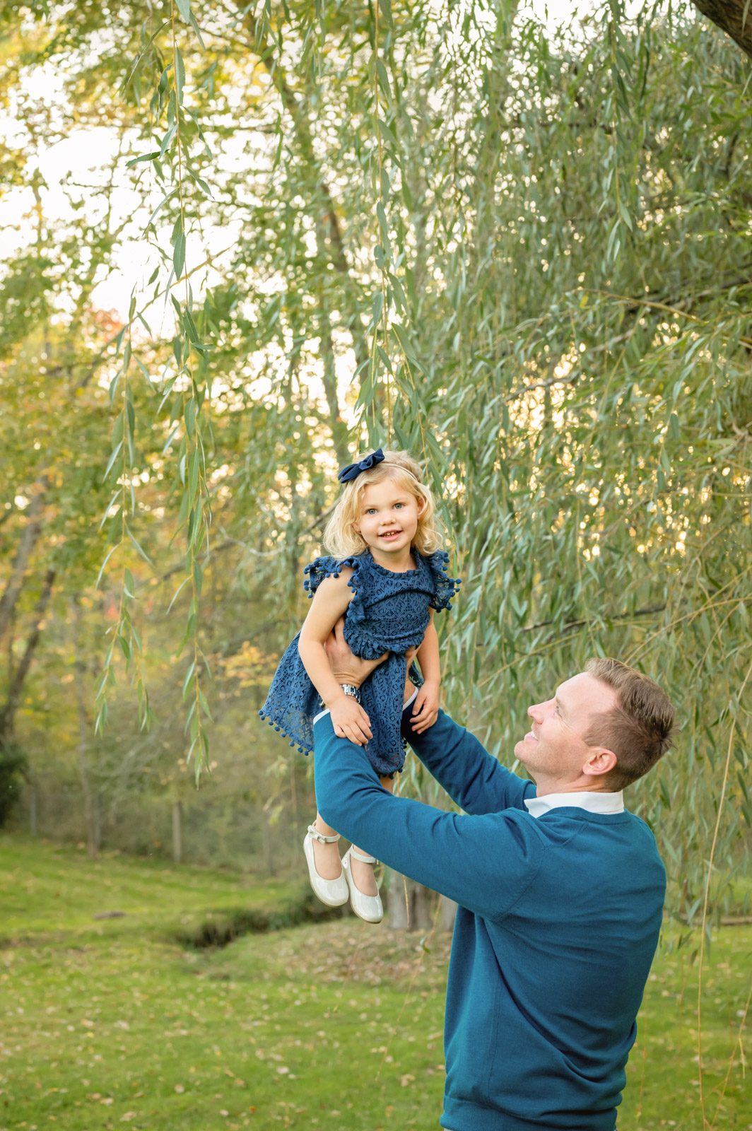 a little girl smiling at the camera as her dad flies her up in the air in front of a willow tree during a family photoshoot