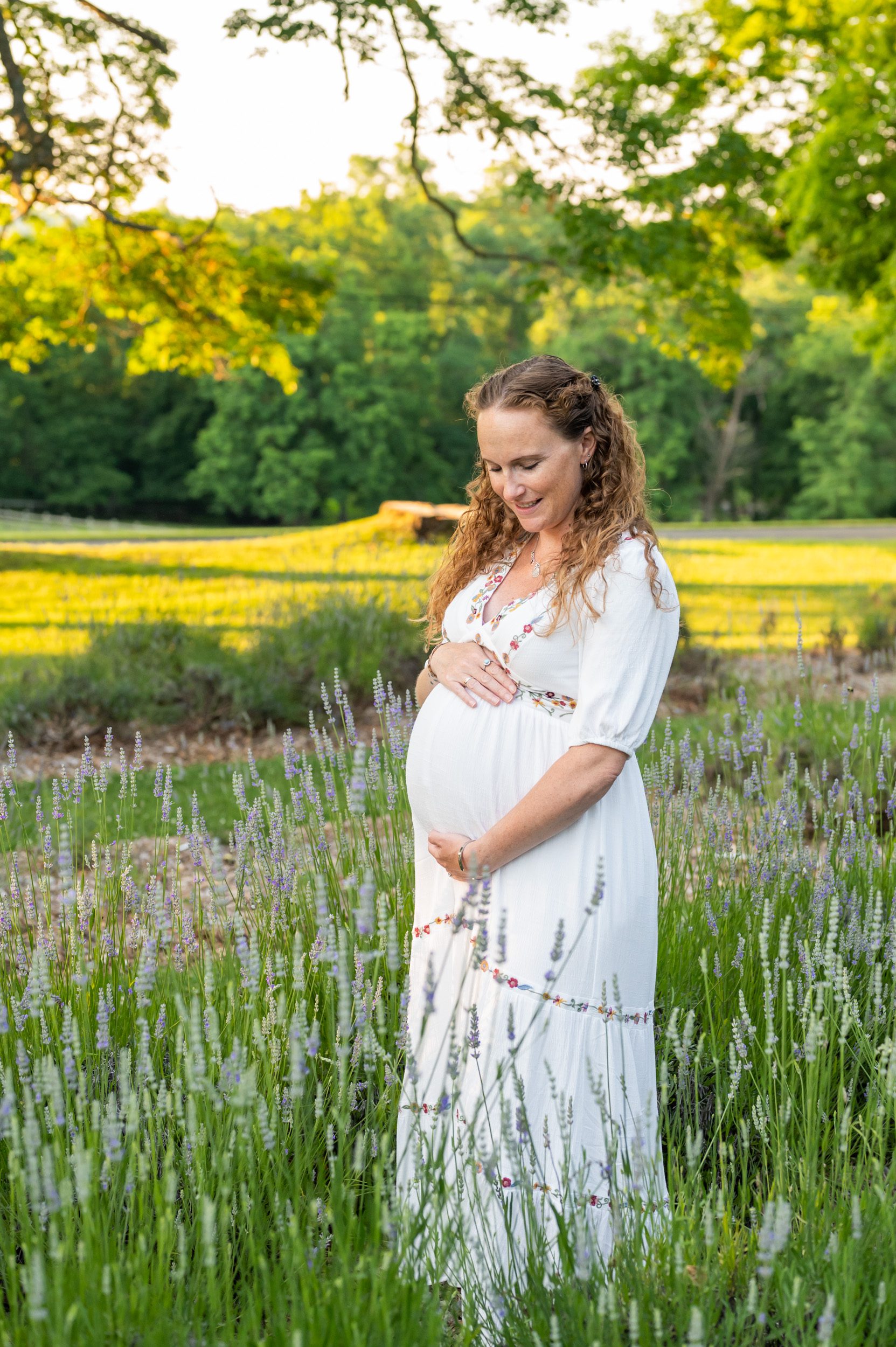 an expecting mother in a white dress with flower embroidery standing in a lavender field as she smiles down at her belly during a maternity photo session