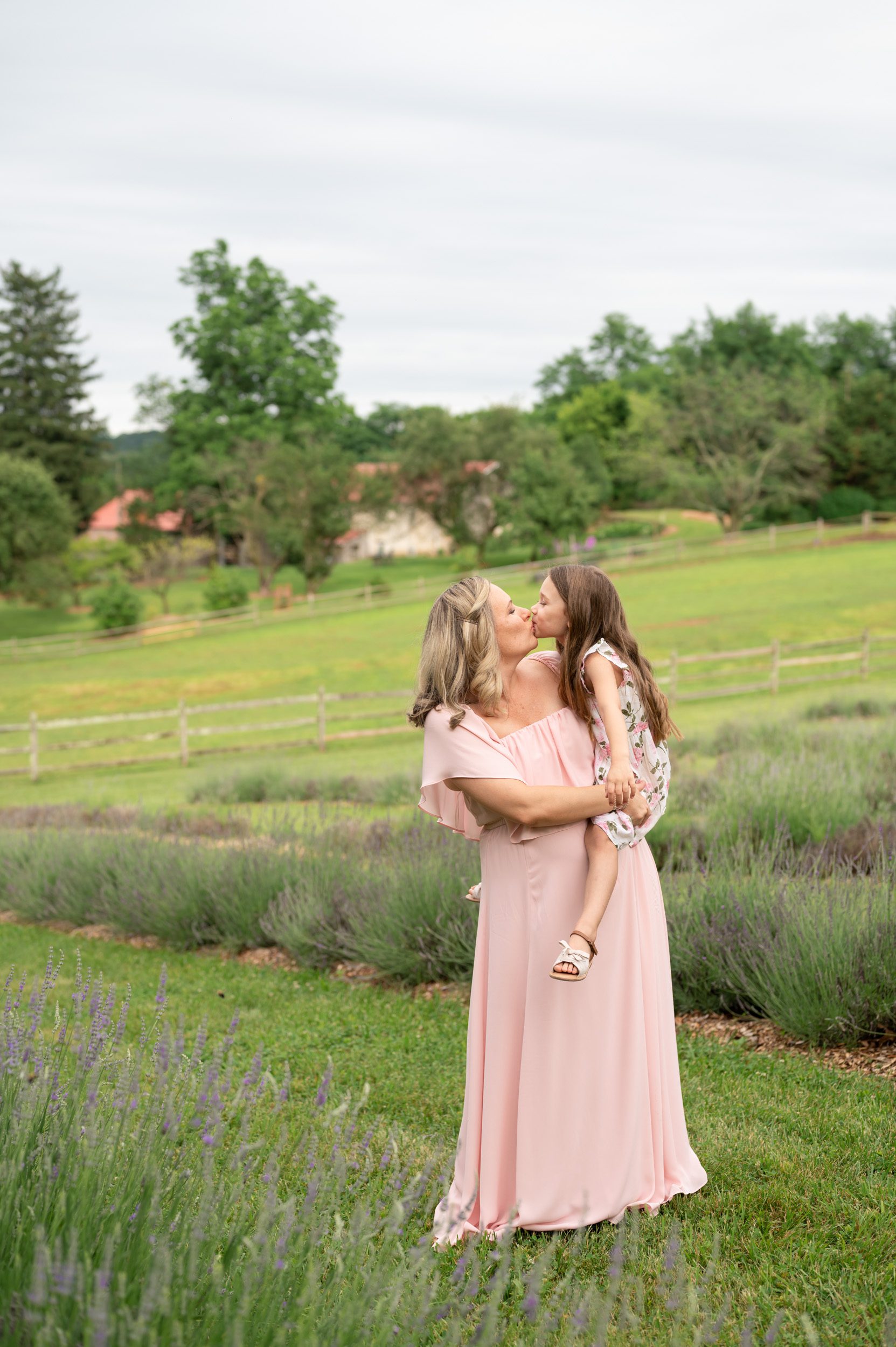 an expecting mom in a pink dress holding her daughter in a field of lavender as they kiss each other during a maternity photoshoot