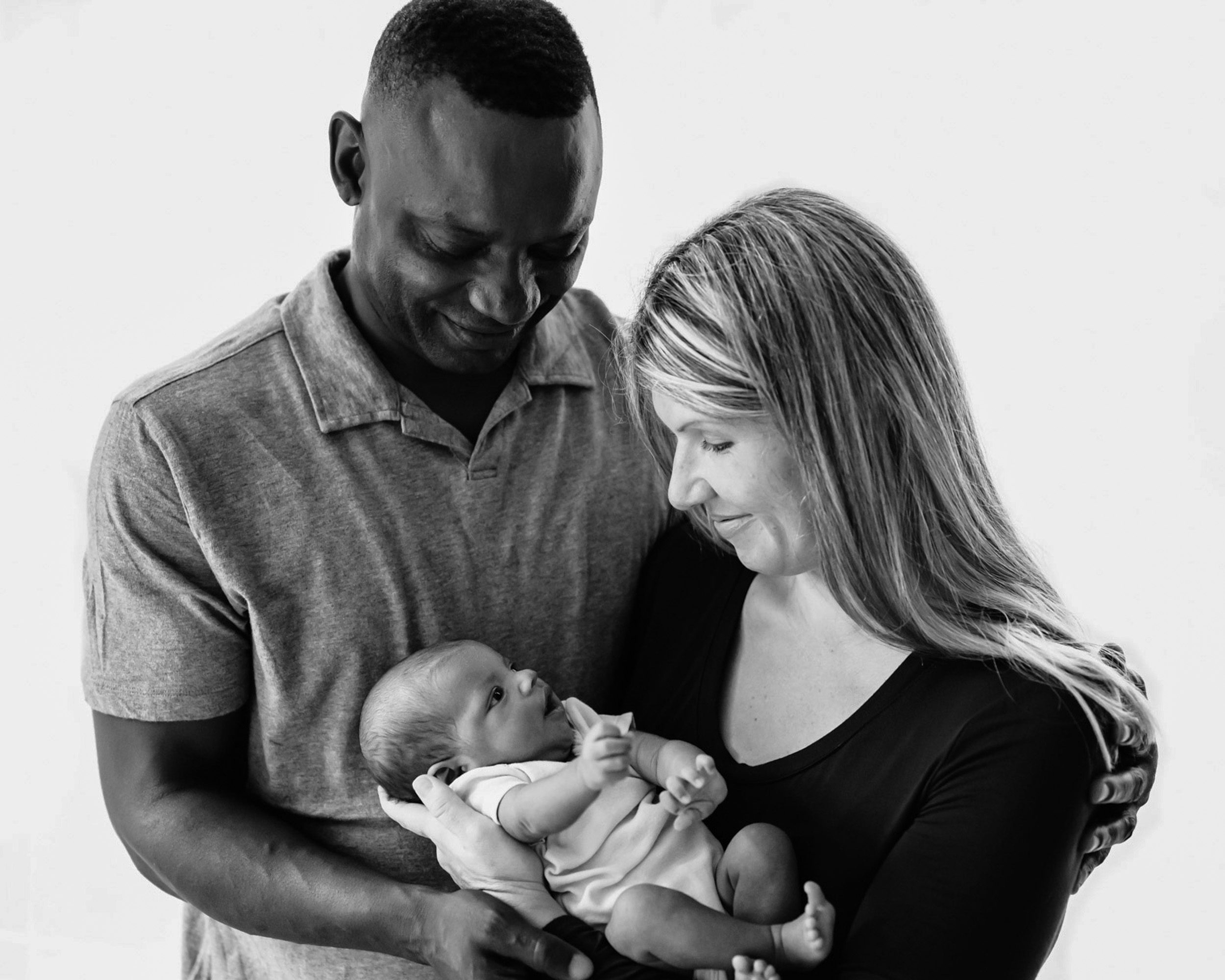 a black and white backlit picture of a mom and dad smiling down at their baby girl as she looks up at mom during a lifestyle newborn photo session