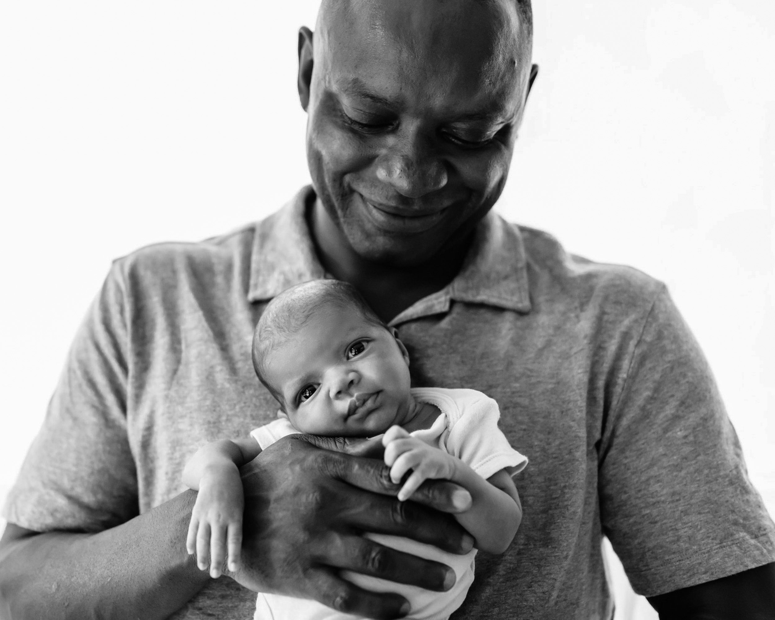 black and white backlit picture of a father holding his newborn baby girl against his chest as she looks toward the camera during an in home newborn photoshoot