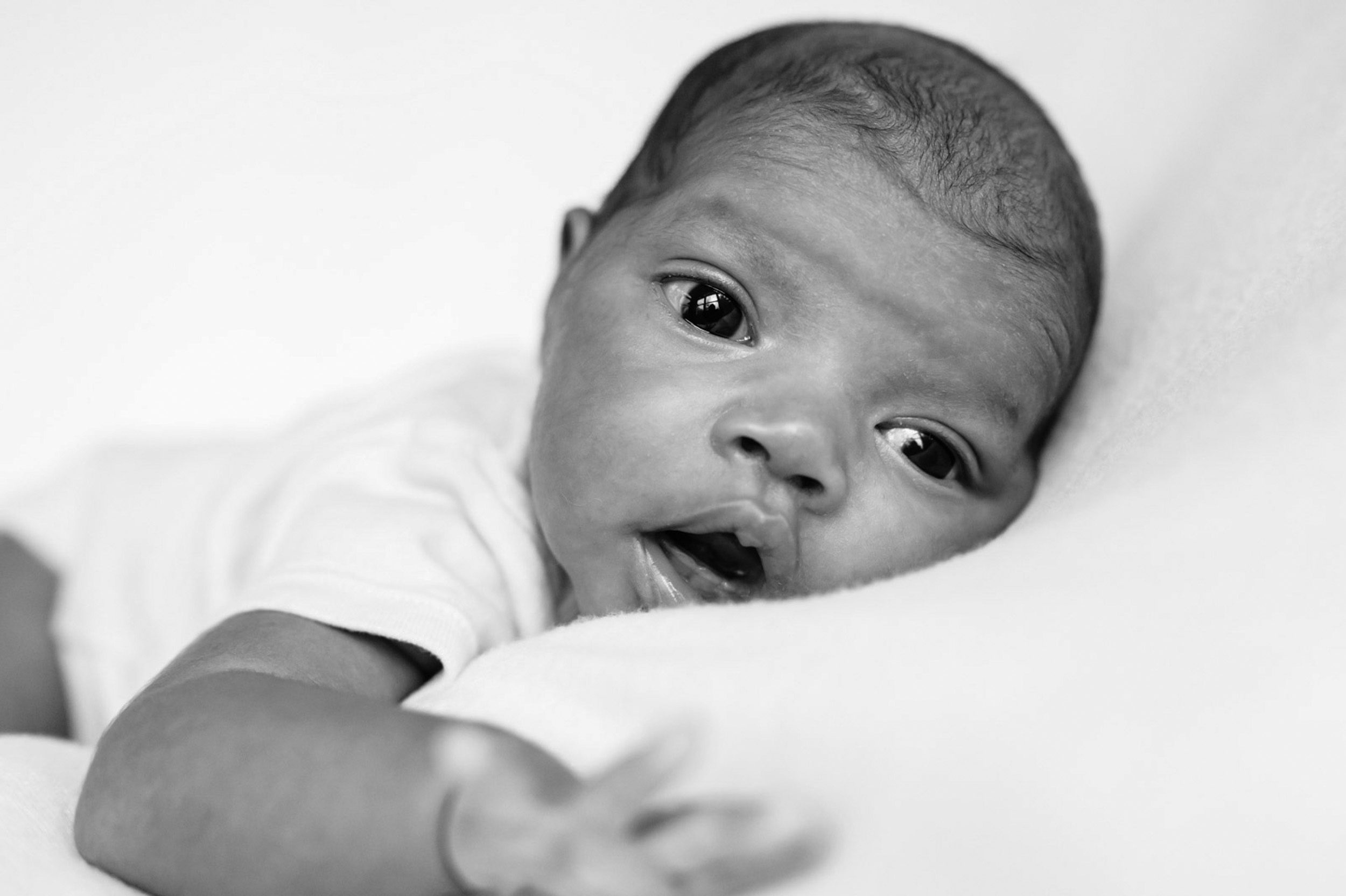 black and white picture of a newborn baby girl laying on her belly on a white backdrop reaching toward the camera with her hand during a newborn photoshoot
