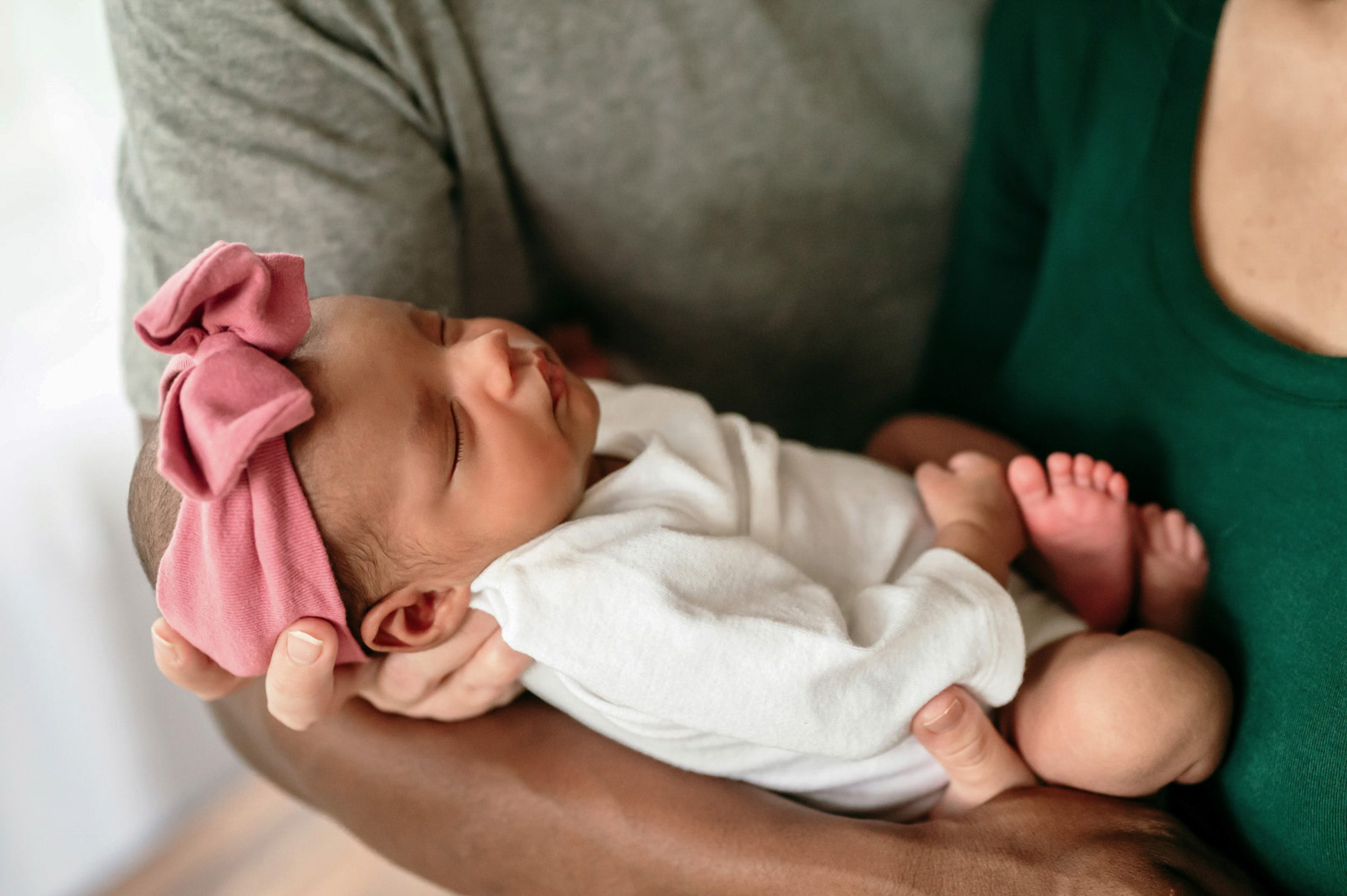 close up picture of a baby girl wearing a white onesie and pink headband nestled in her parents arms during a lifestyle newborn photoshoot