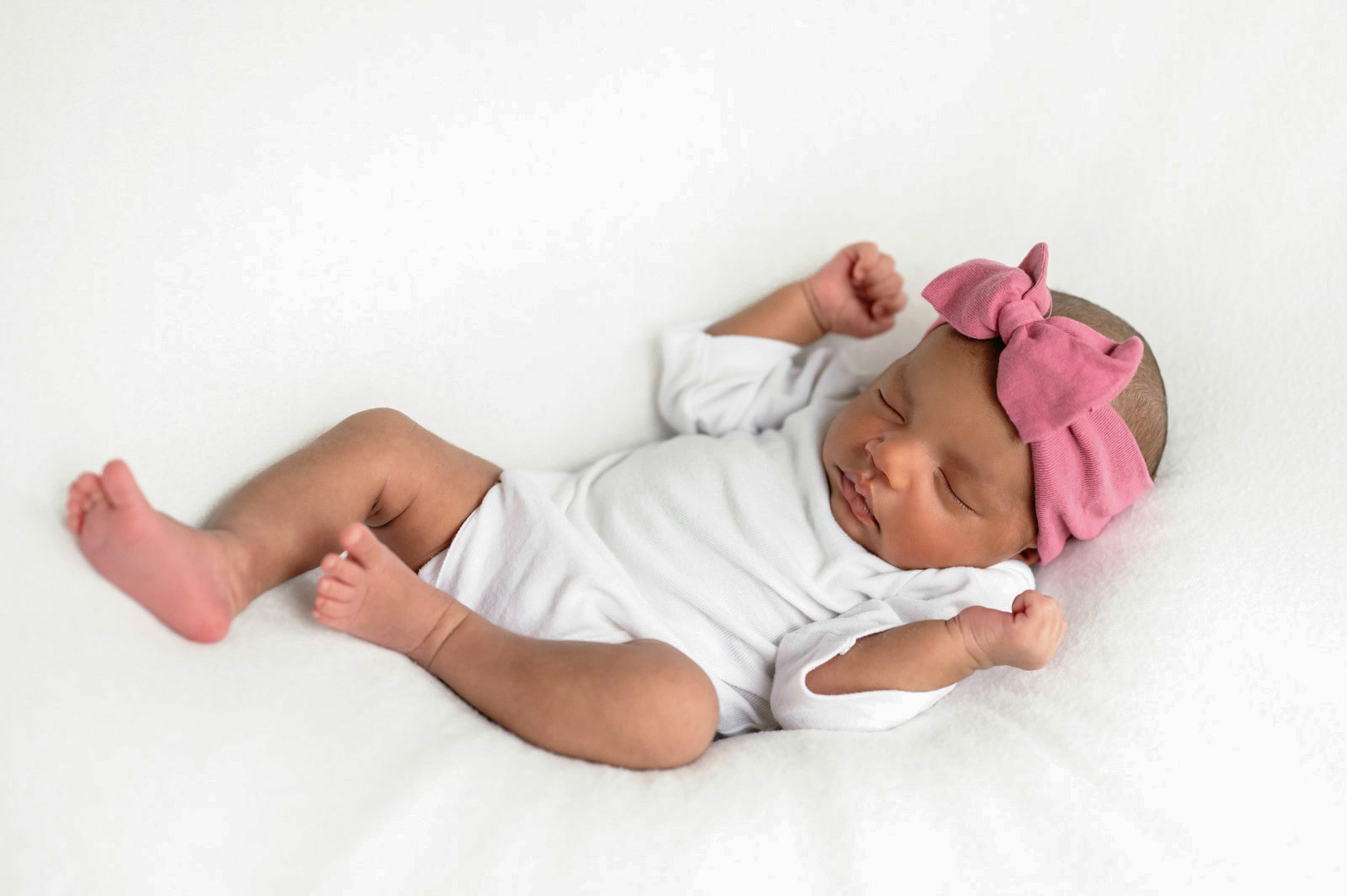 a baby girl wearing a white onesie and pink handband laying on a white backdrop during a newborn photoshoot
