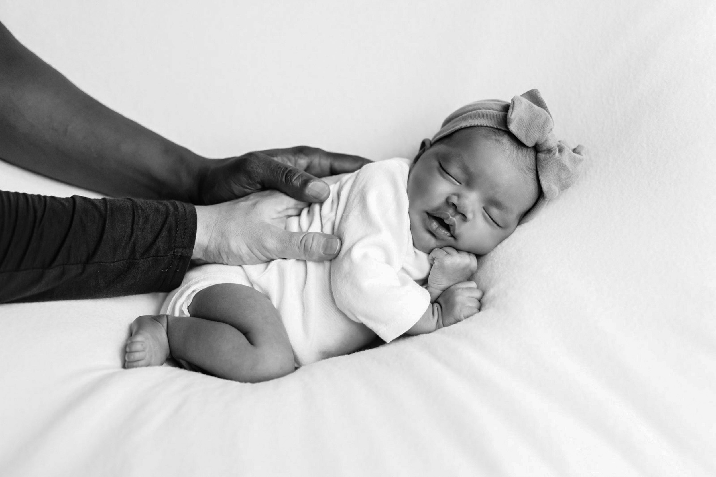 a black and white picture of a baby girl in a white onesie laying on a white backdrop with both her parents resting their hands on her back during a newborn photoshoot