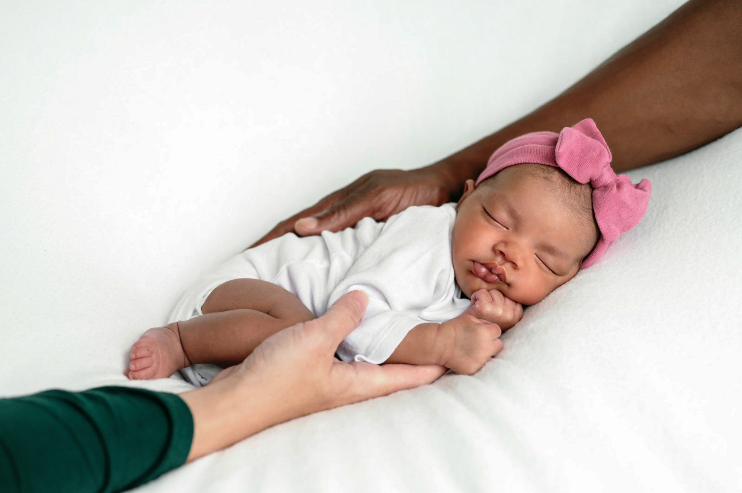 a baby girl in a white onesie and pink headband laying on her stomach on a white backdrop while her mom and dad cradle her with their arms during a newborn photo session