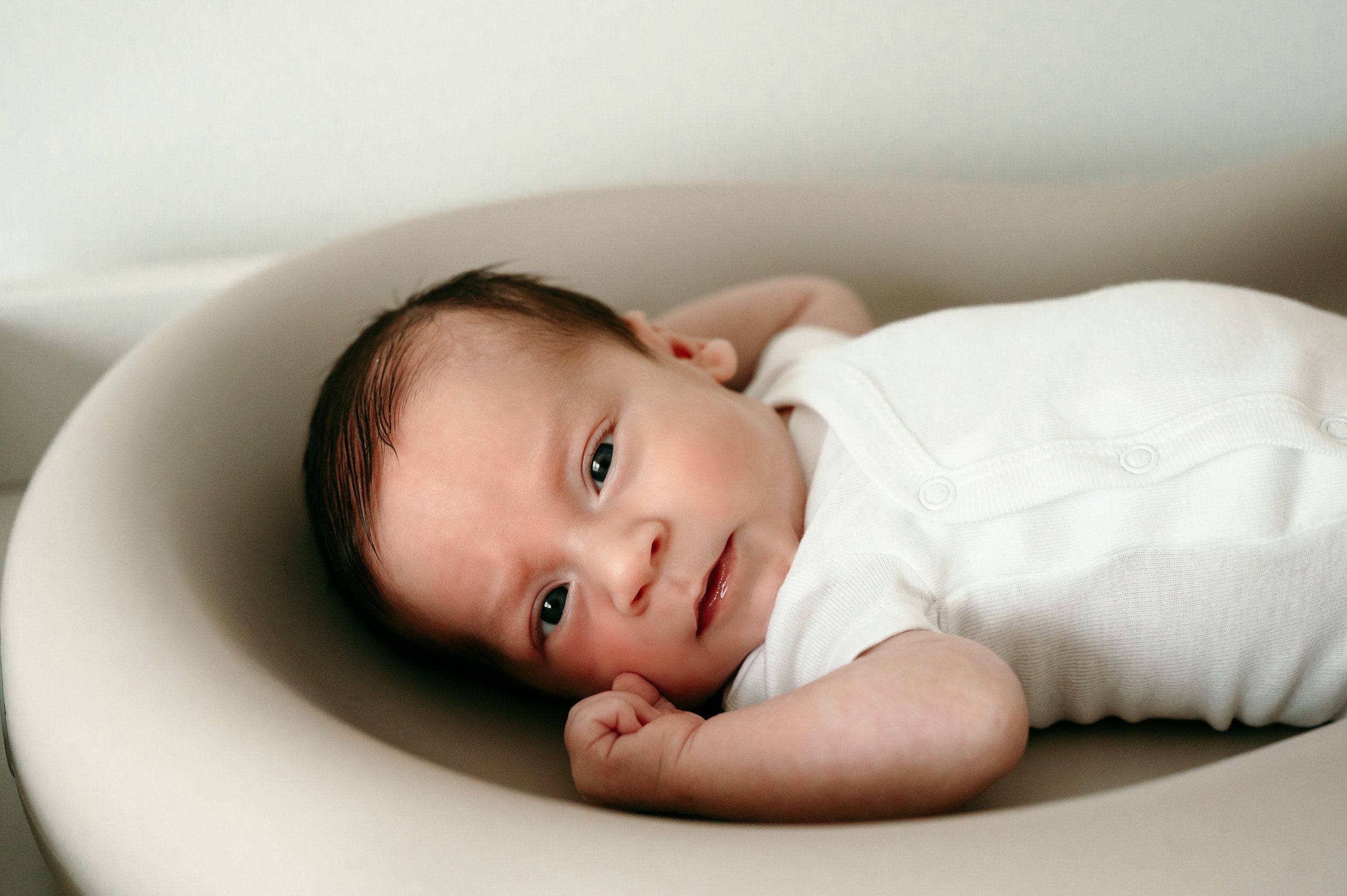 a baby boy in a white onesie laying on his changing table and looking at the camera during a newborn photography session