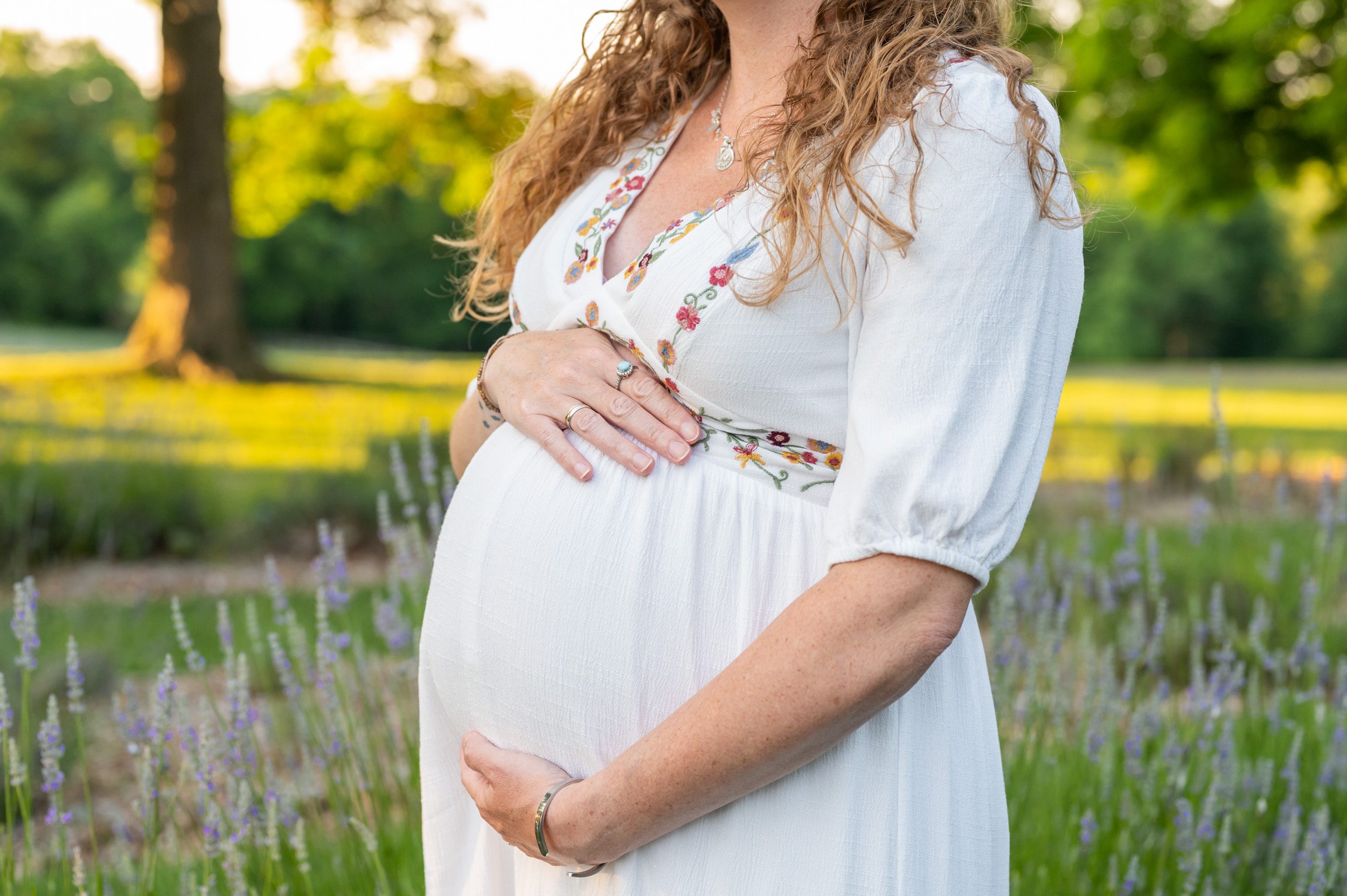 a close up picture of a pregnant mom in a white dress with flower embroidery standing in a lavender field and cradling her belly during a maternity photo session