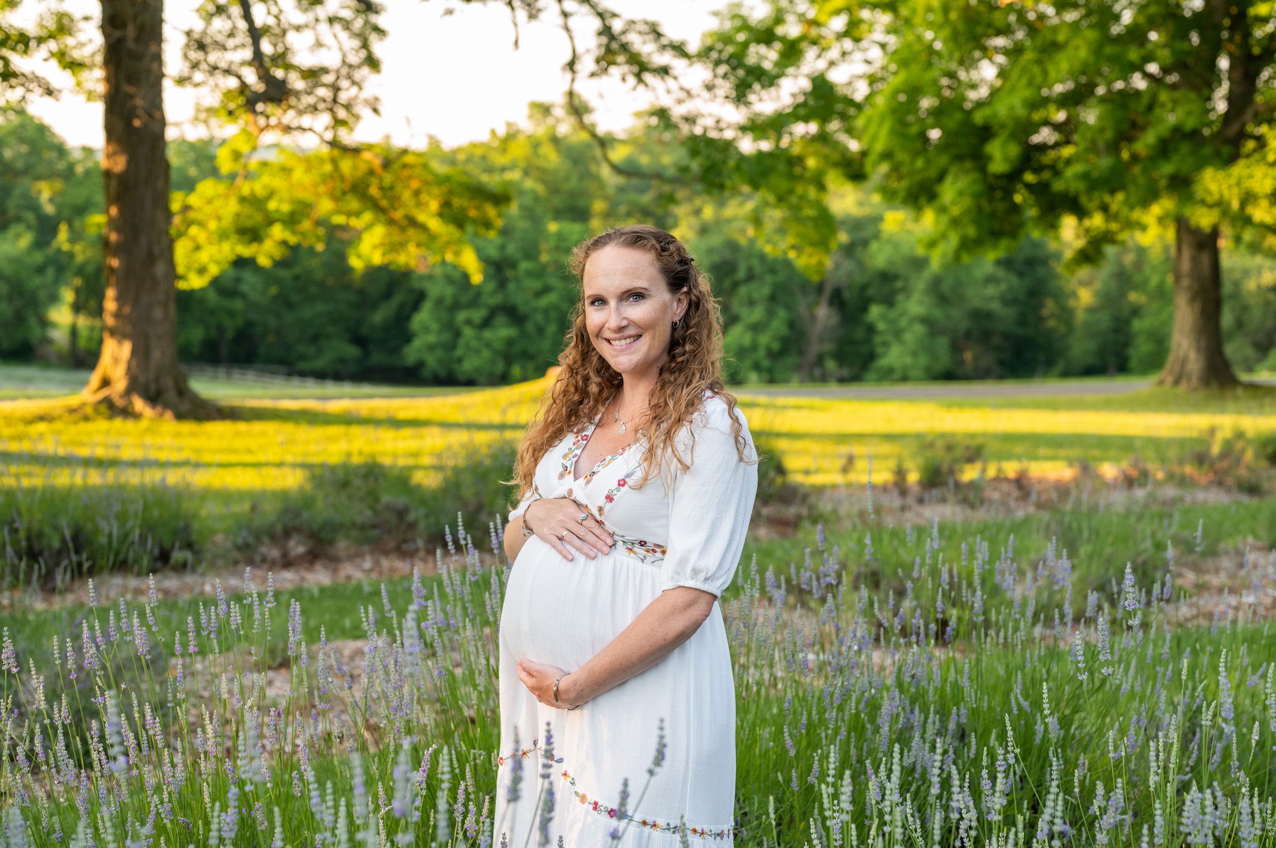 a pregnant mom standing in a lavender field and smiling at the camera as she cradles her belly during a maternity photoshoot