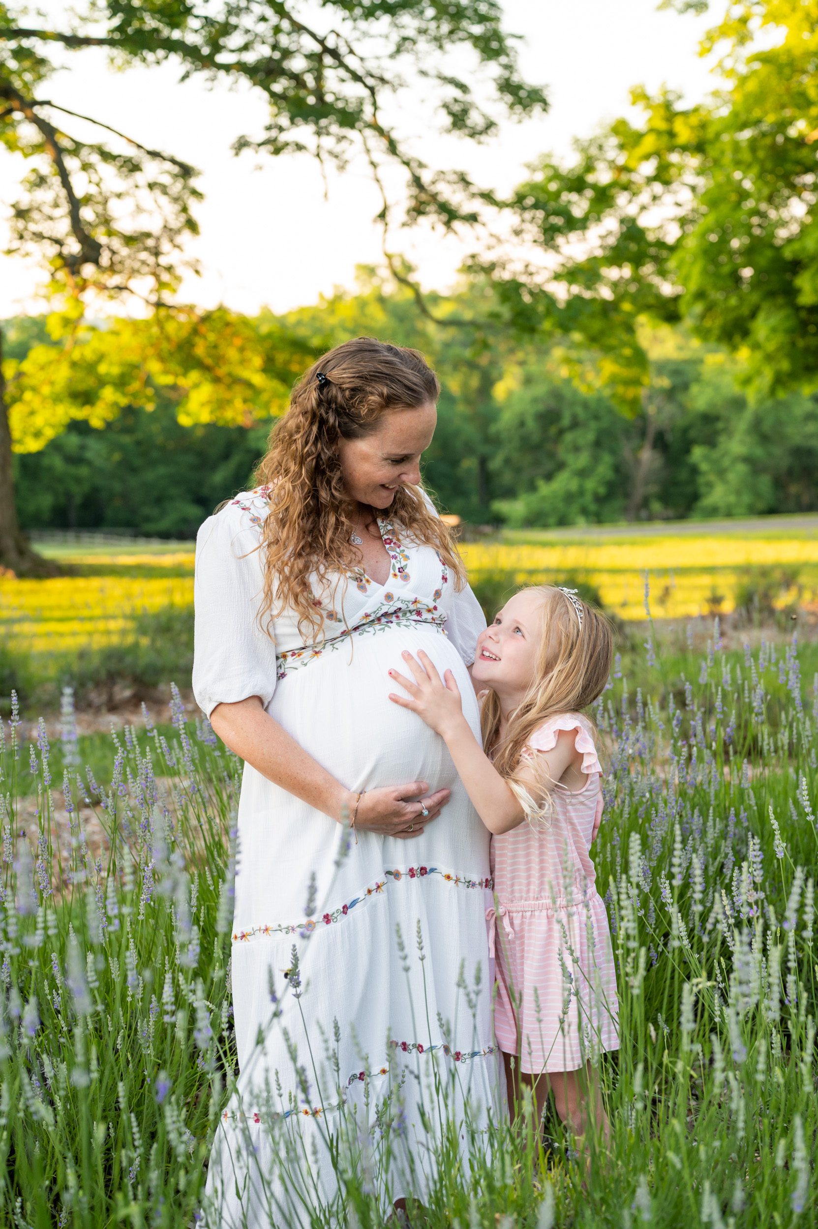 a young girl smiling up at her pregnant mom and touching her belly during a maternity photoshoot