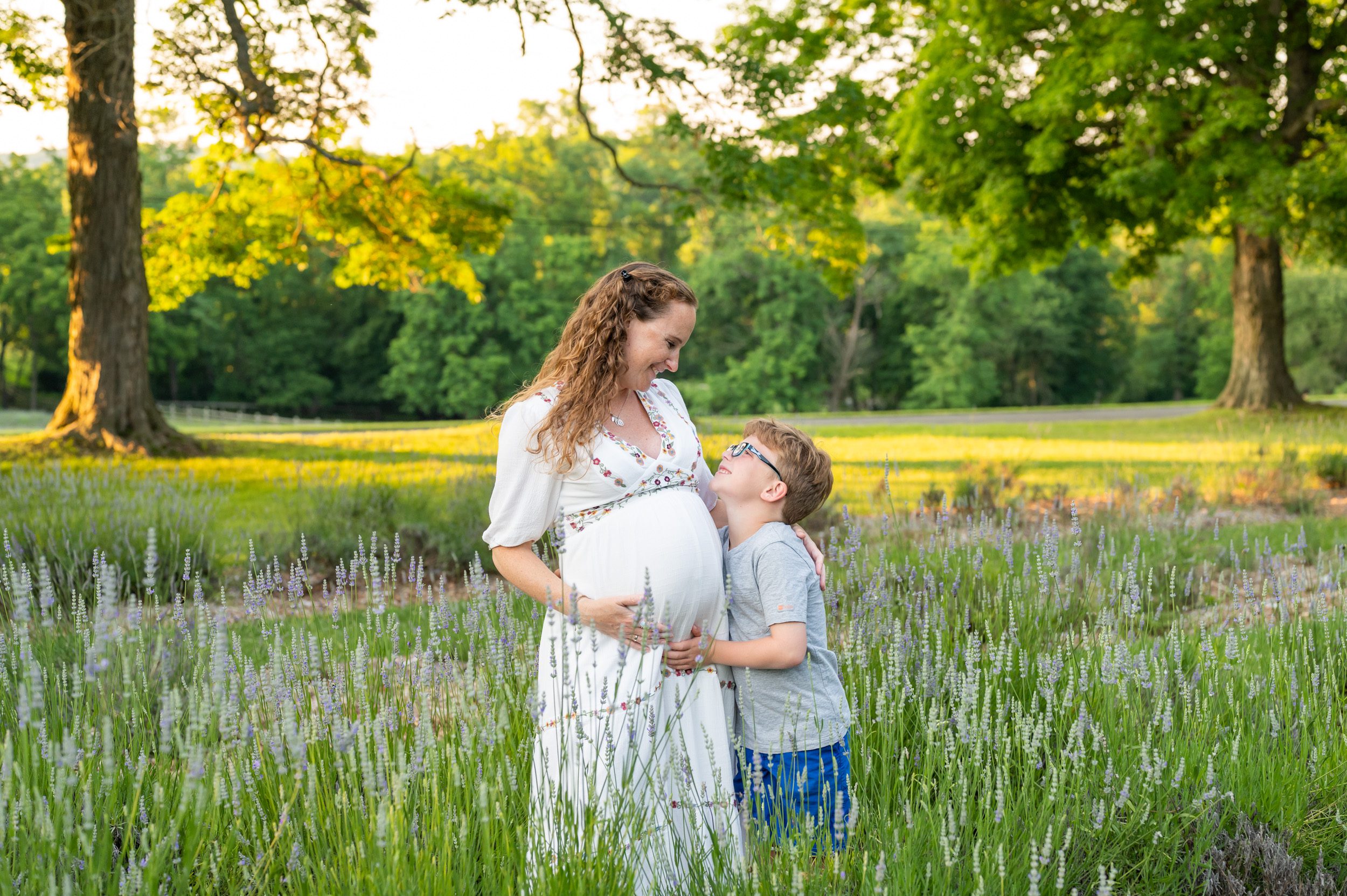 a pregnant mom standing in a lavender field with her young son as they both smile at each other during a maternity photoshoot