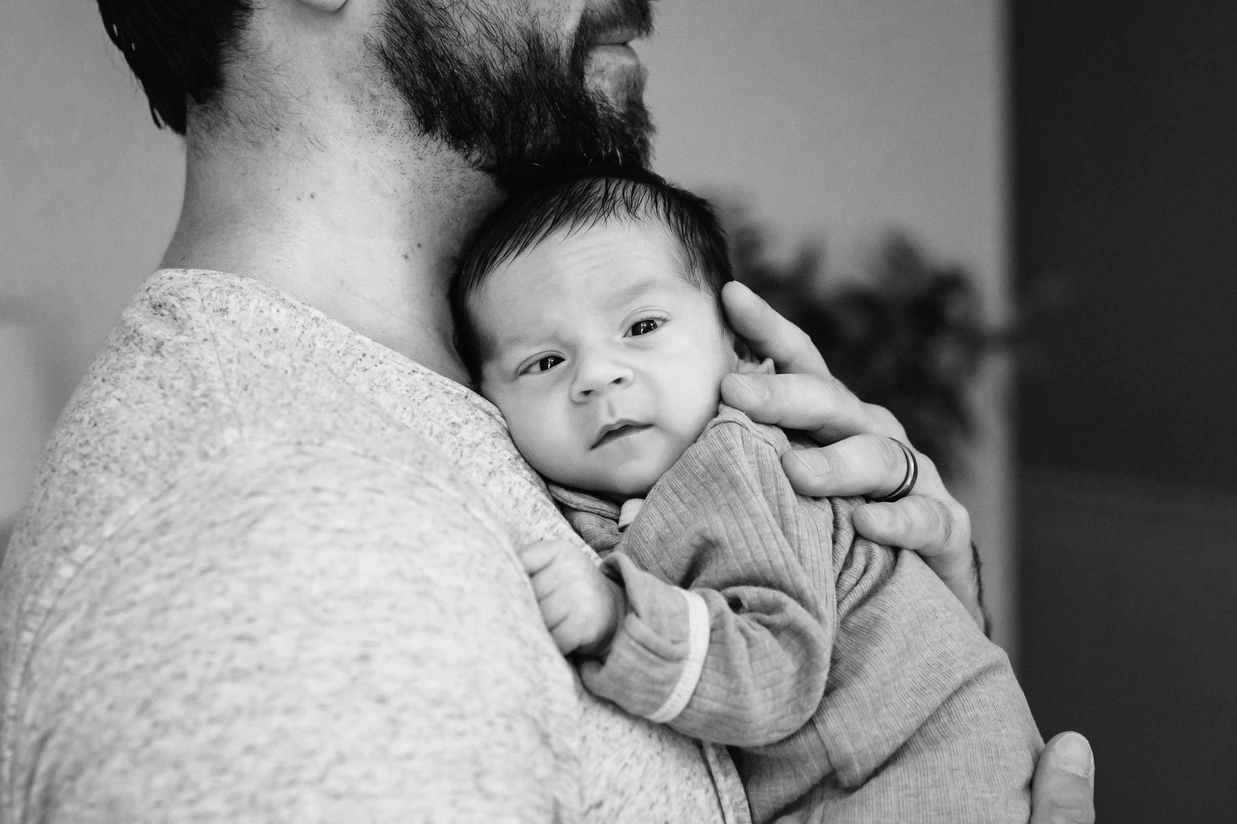 a black and white picture of a newborn baby boy snuggled up against his dad's chest and looking at the camera during a newborn photo session
