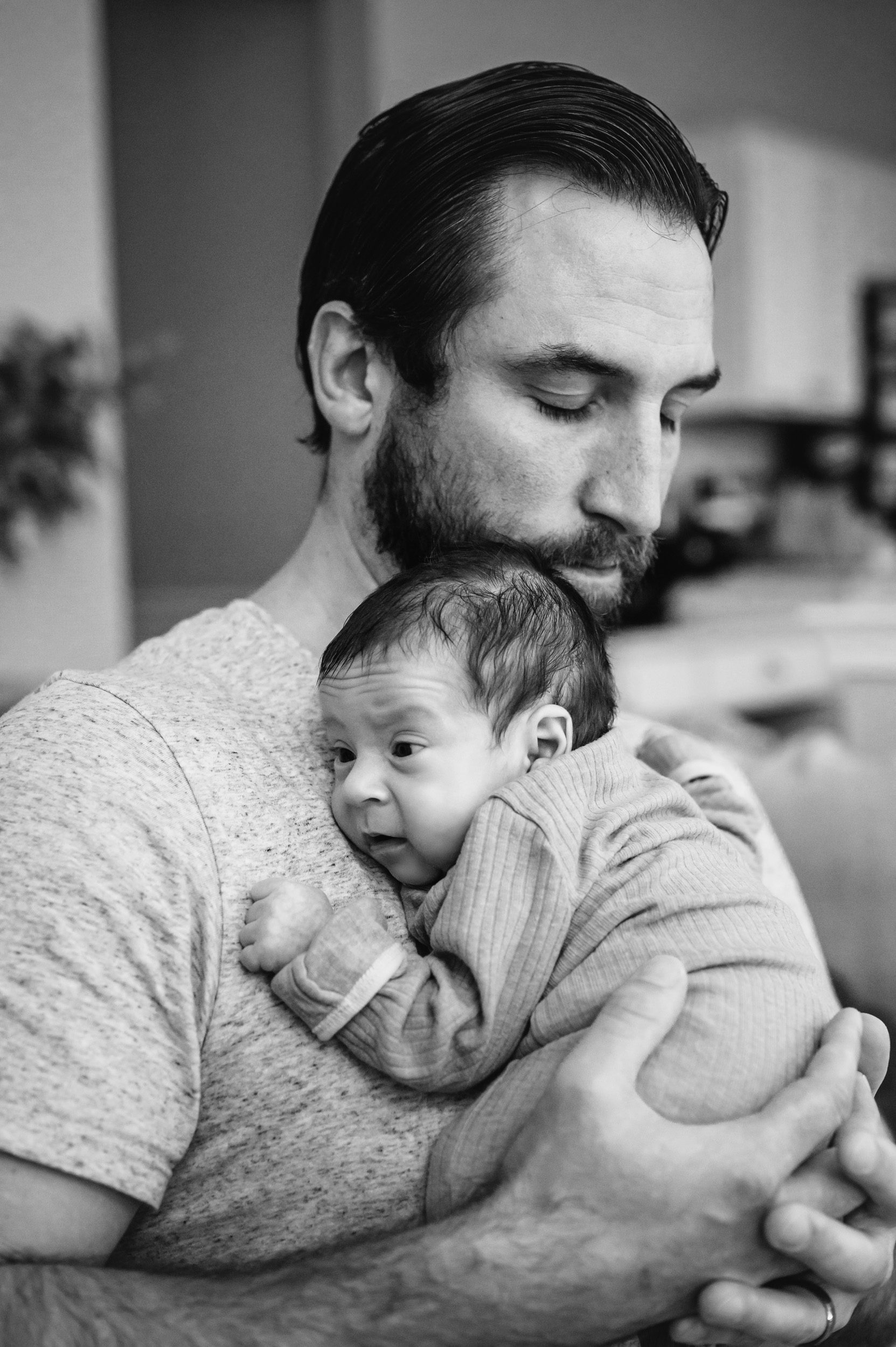 a black and white picture of a new dad holding his baby boy against his chest during a newborn photoshoot