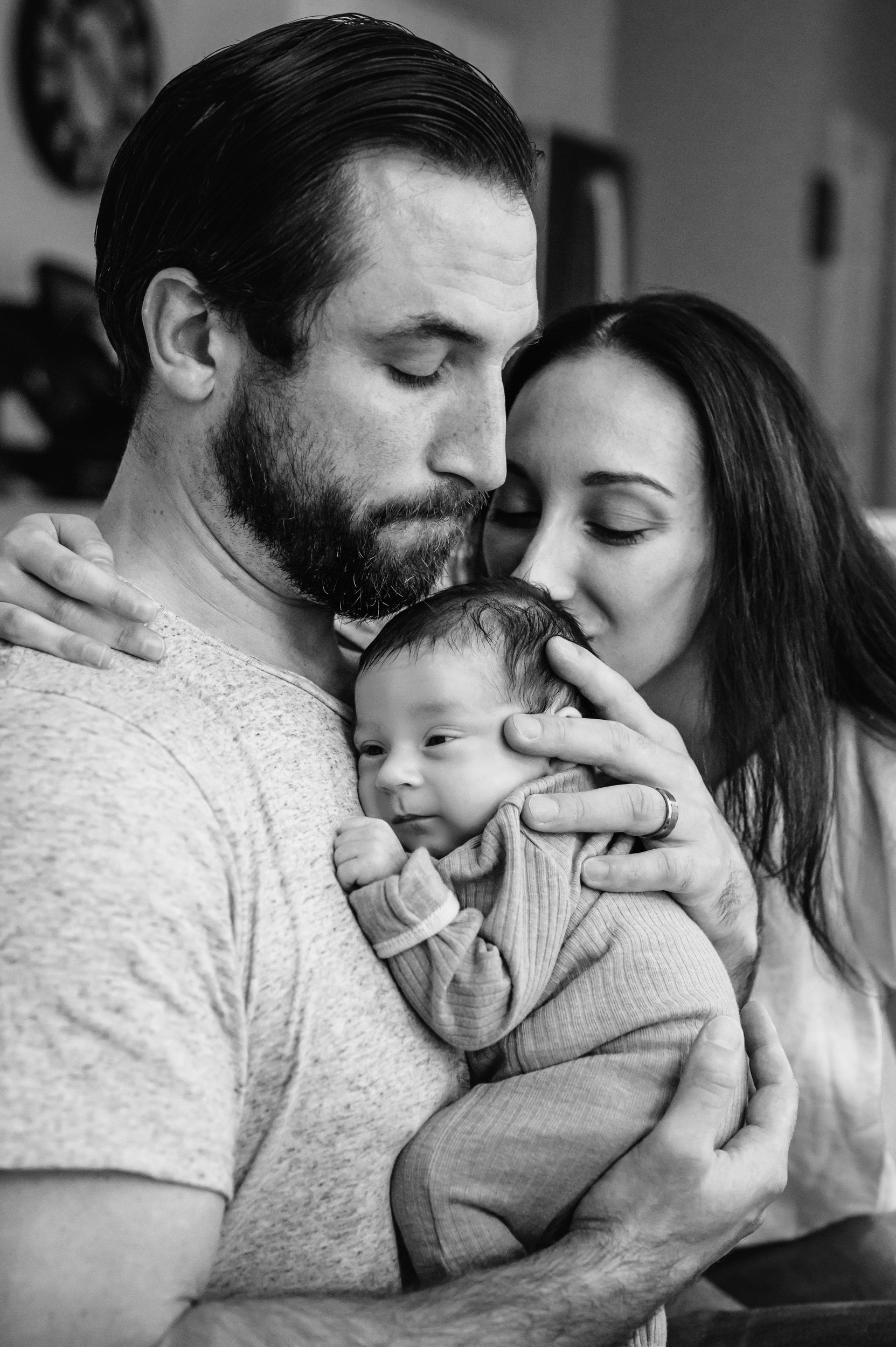 a black and white picture of a new dad holding his baby boy against his chest while mom kisses the baby on the back of the head during a newborn photo session