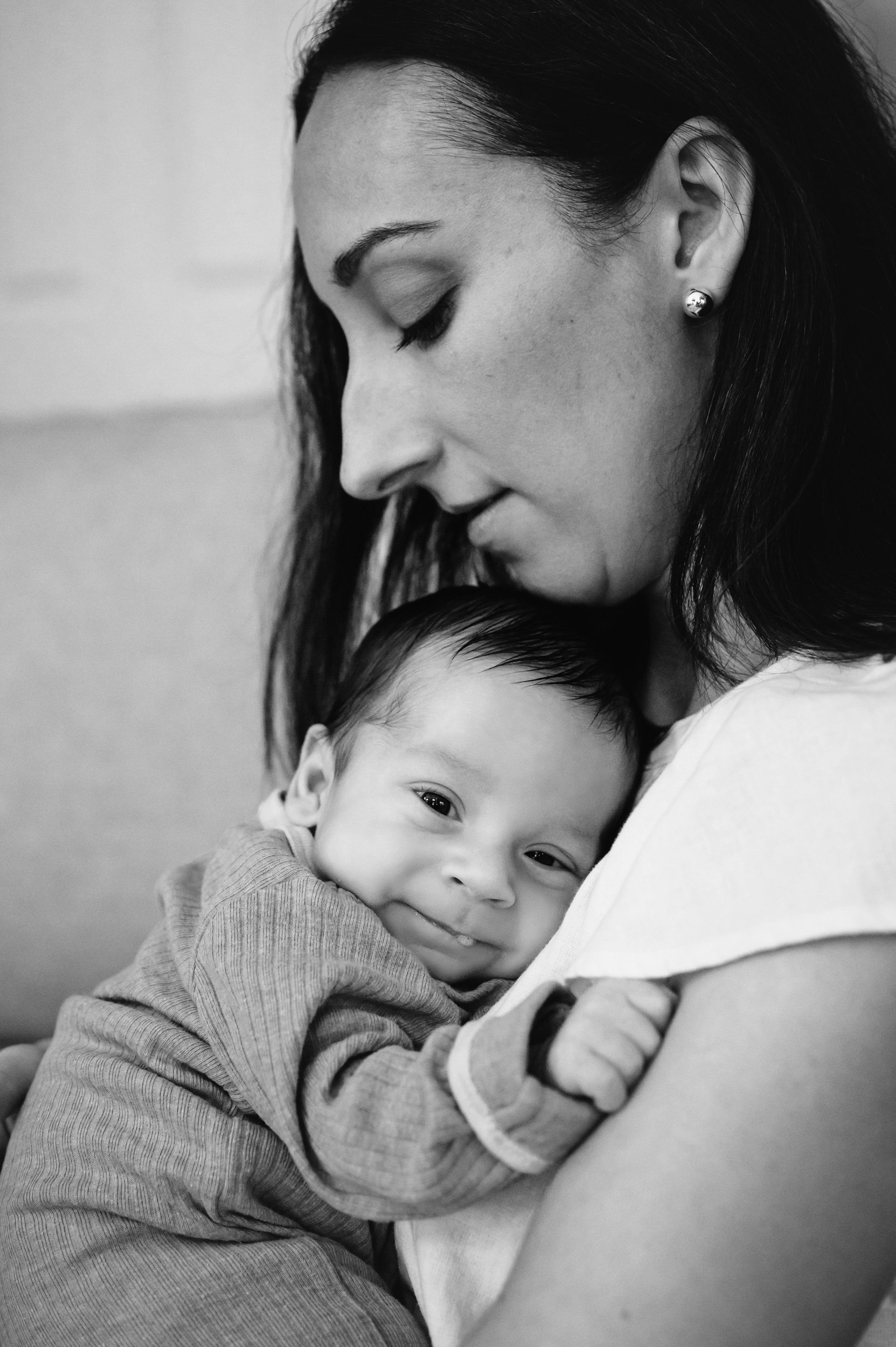 a black and white picture of a new mom holding her baby boy against her chest as he looks at the camera and blows a spit bubble during a newborn photo session