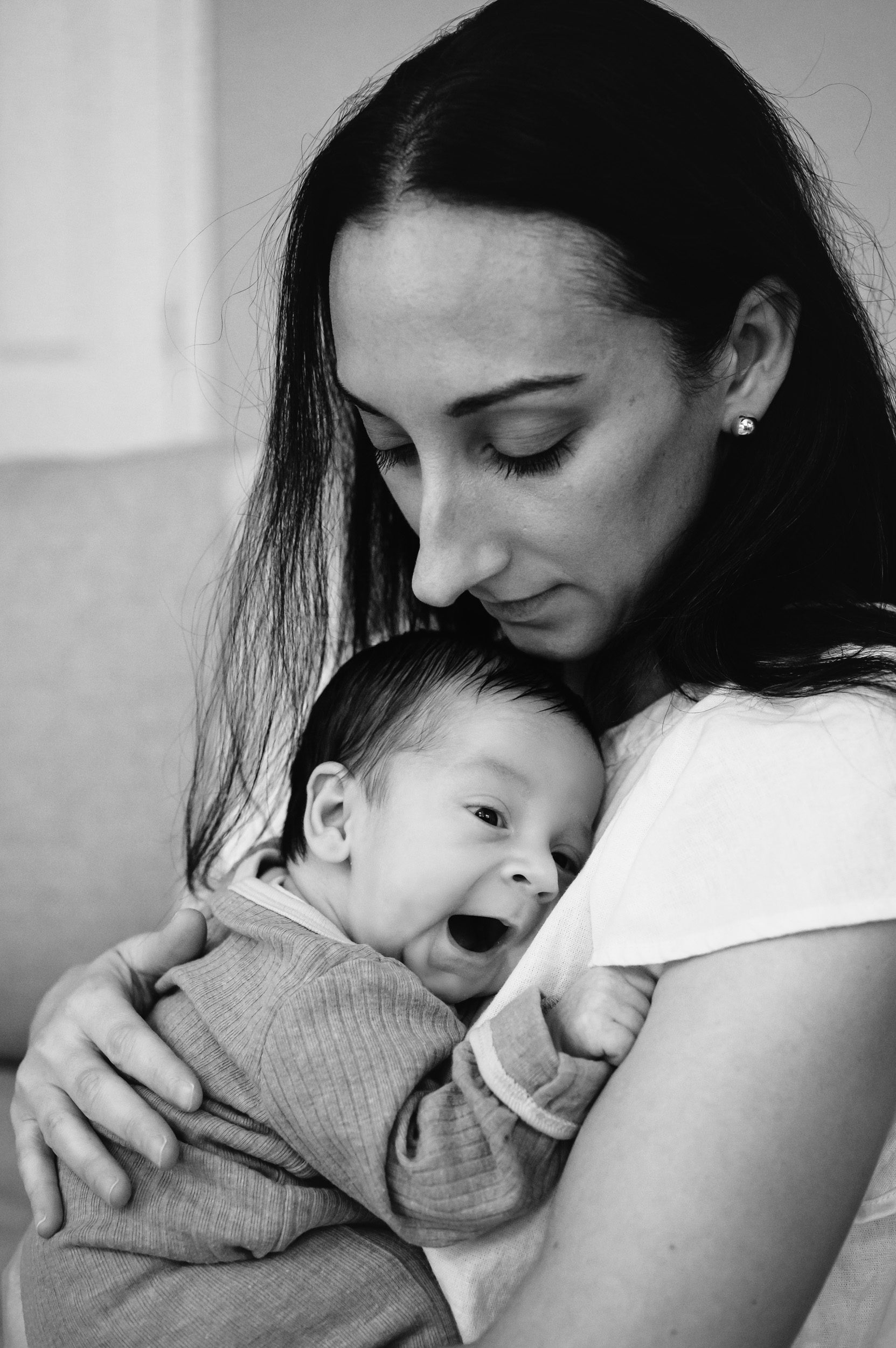 a black and white picture of a newborn baby boy snuggled against his mom's chest and yawning during an in home newborn photoshoot