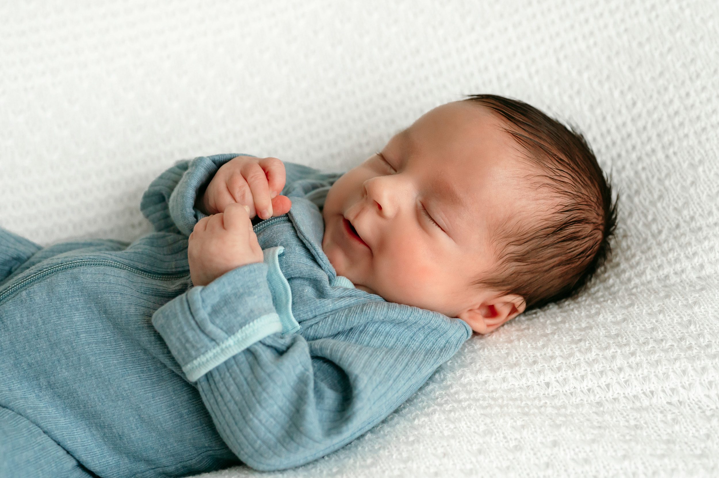 a newborn baby boy in a blue sleeper laying on a white backdrop during a newborn photoshoot