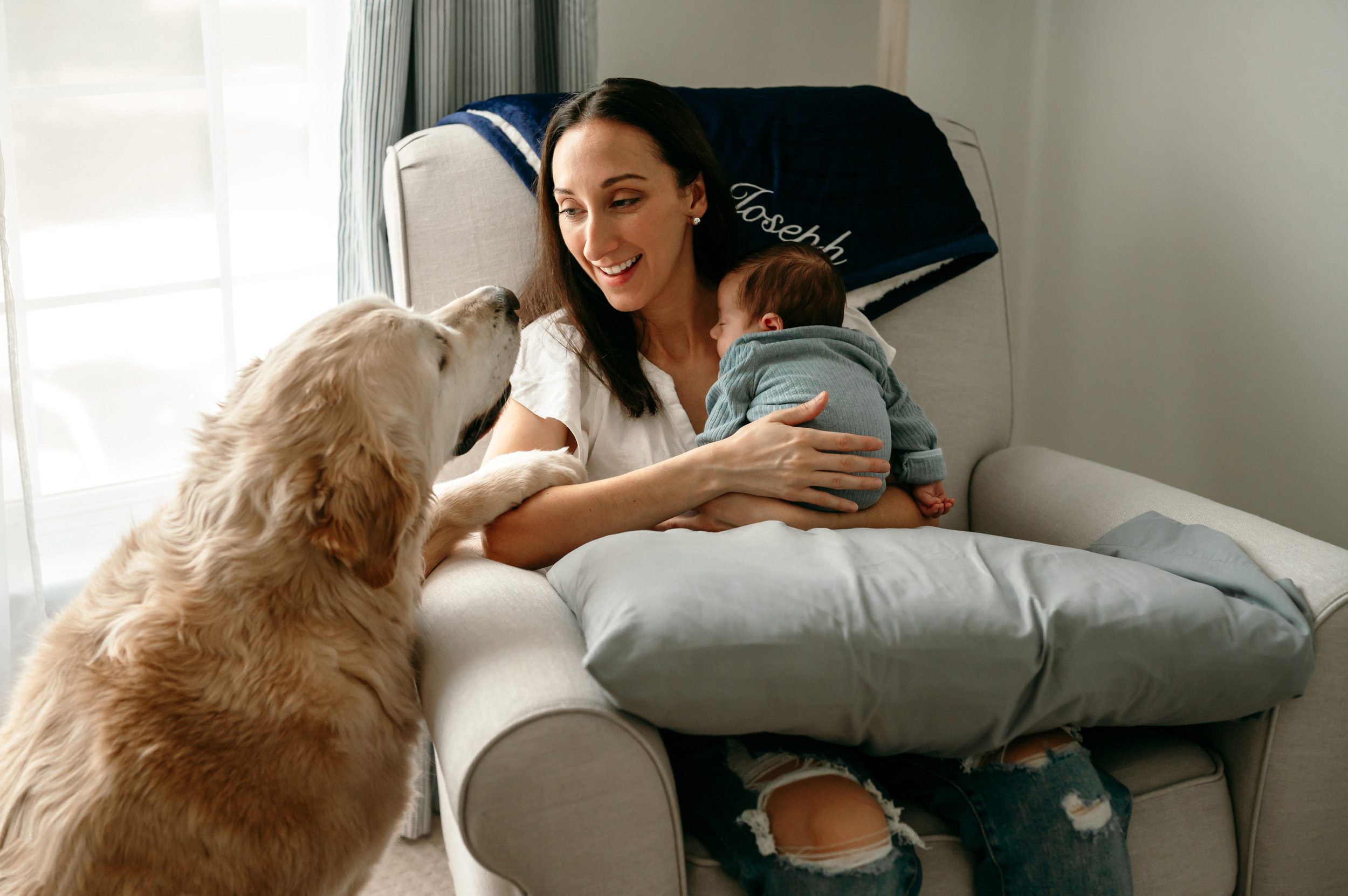 a new mom sitting in a rocking chair with her baby boy against her chest and smiling at her dog as he rests his paw on her arm during an in home newborn photoshoot