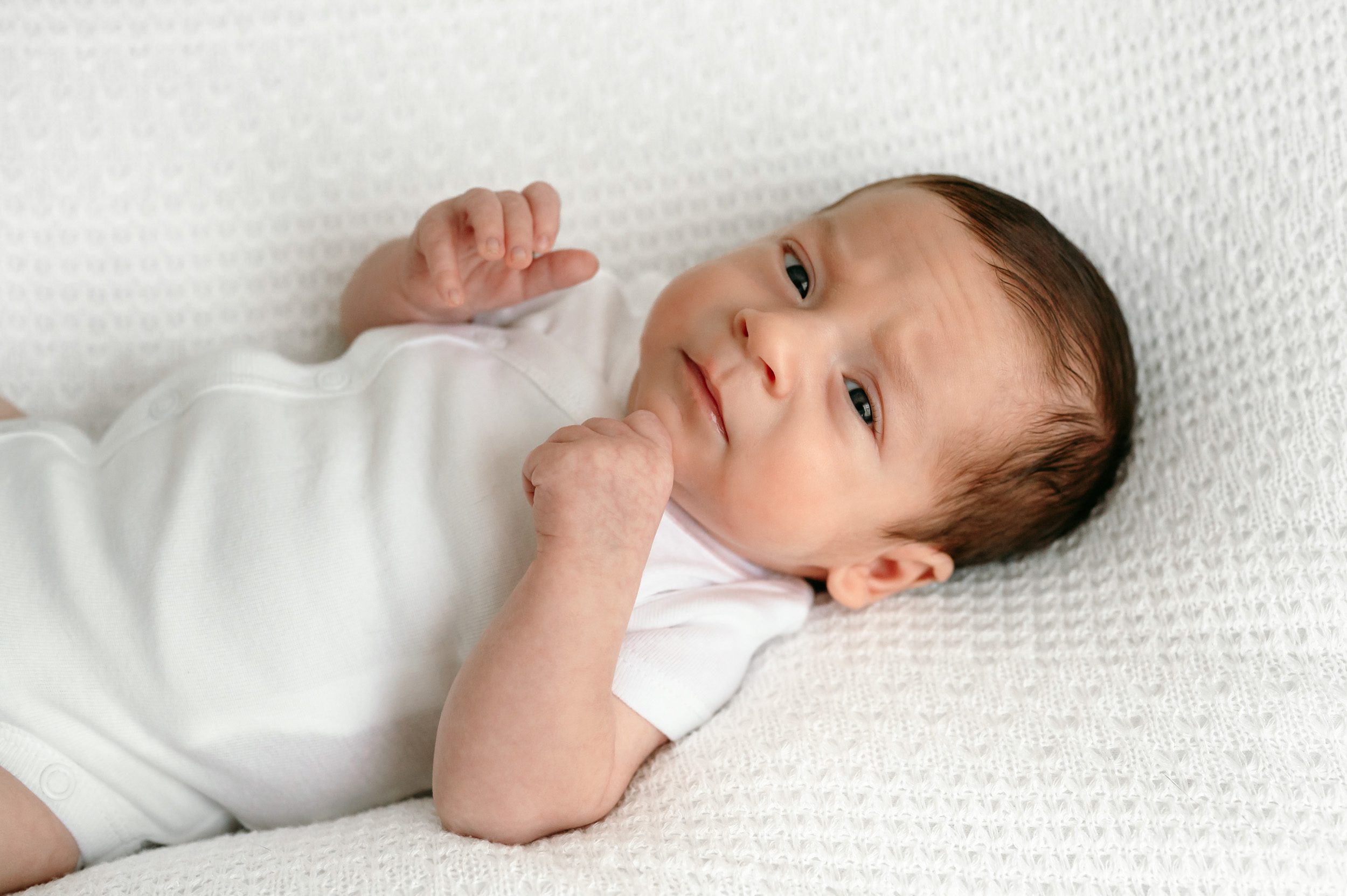 a baby boy in a white onesie on a white backdrop touching his chin and looking at the camera during a newborn photoshoot