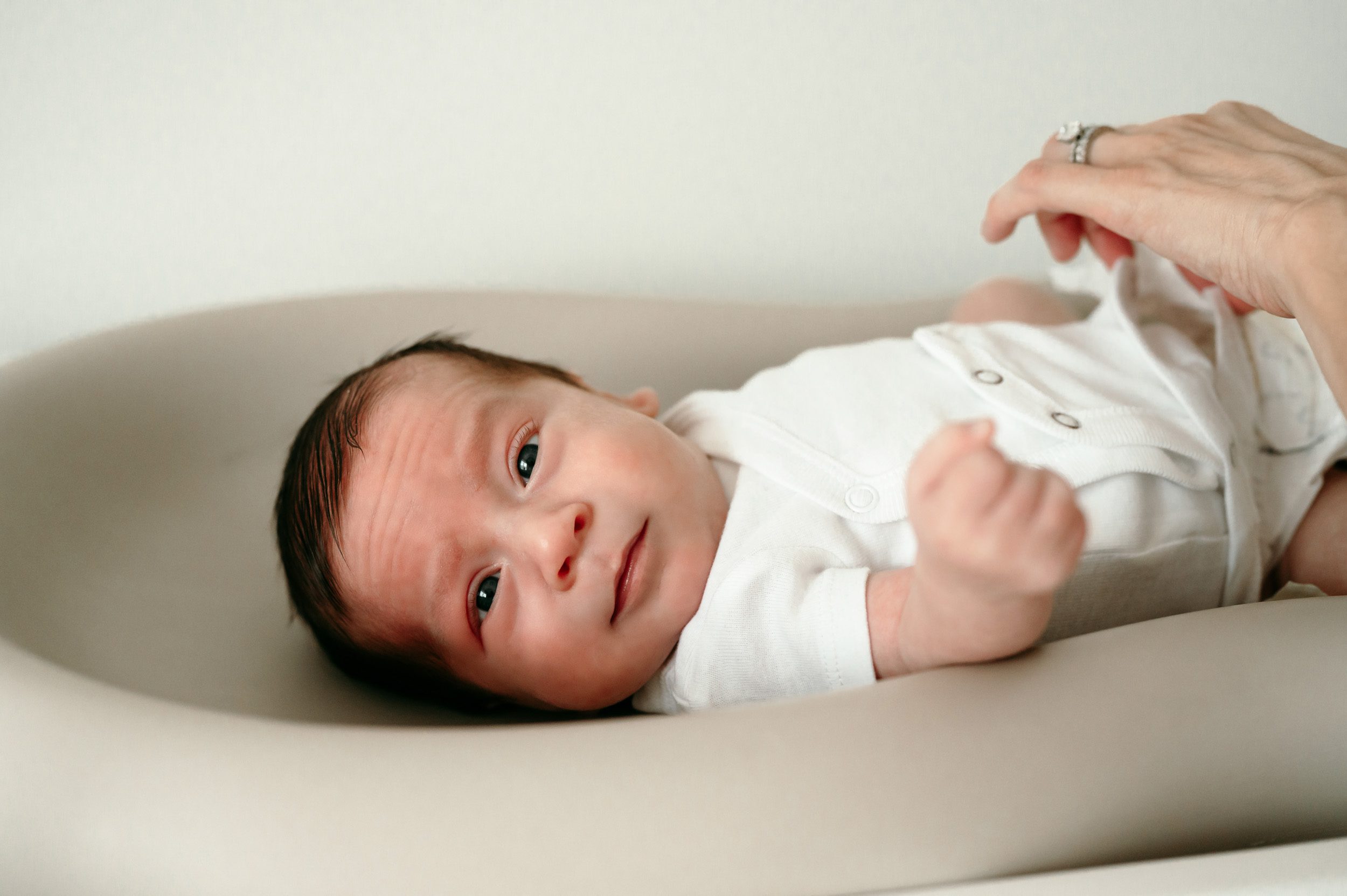 a baby boy in a white onesie laying on his changing table and smiling at the camera as his mom changes his diaper during an in home newborn photoshoot