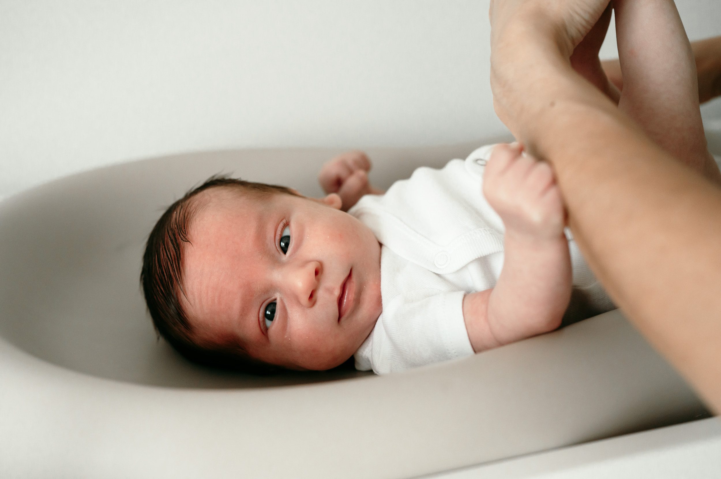 a baby boy in a white onesie laying on his changing table and crossing his eyes at the camera as his mom changes his diaper during an in home newborn photo session