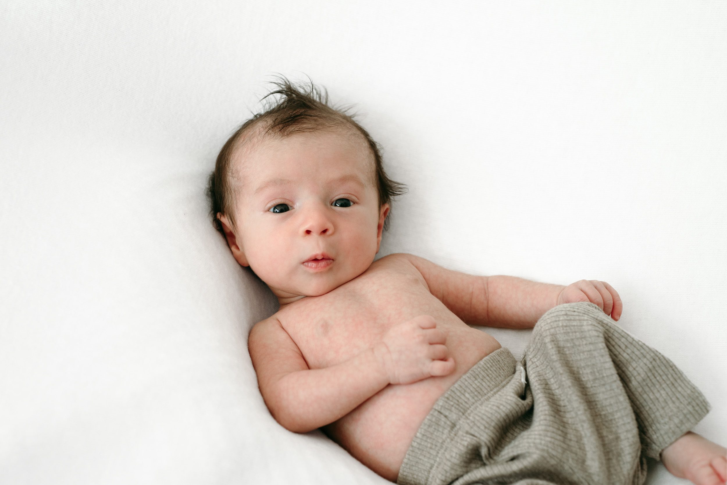 a baby boy wearing taupe pants and laying on a white backdrop looking intently at the camera during a natural newborn photo session