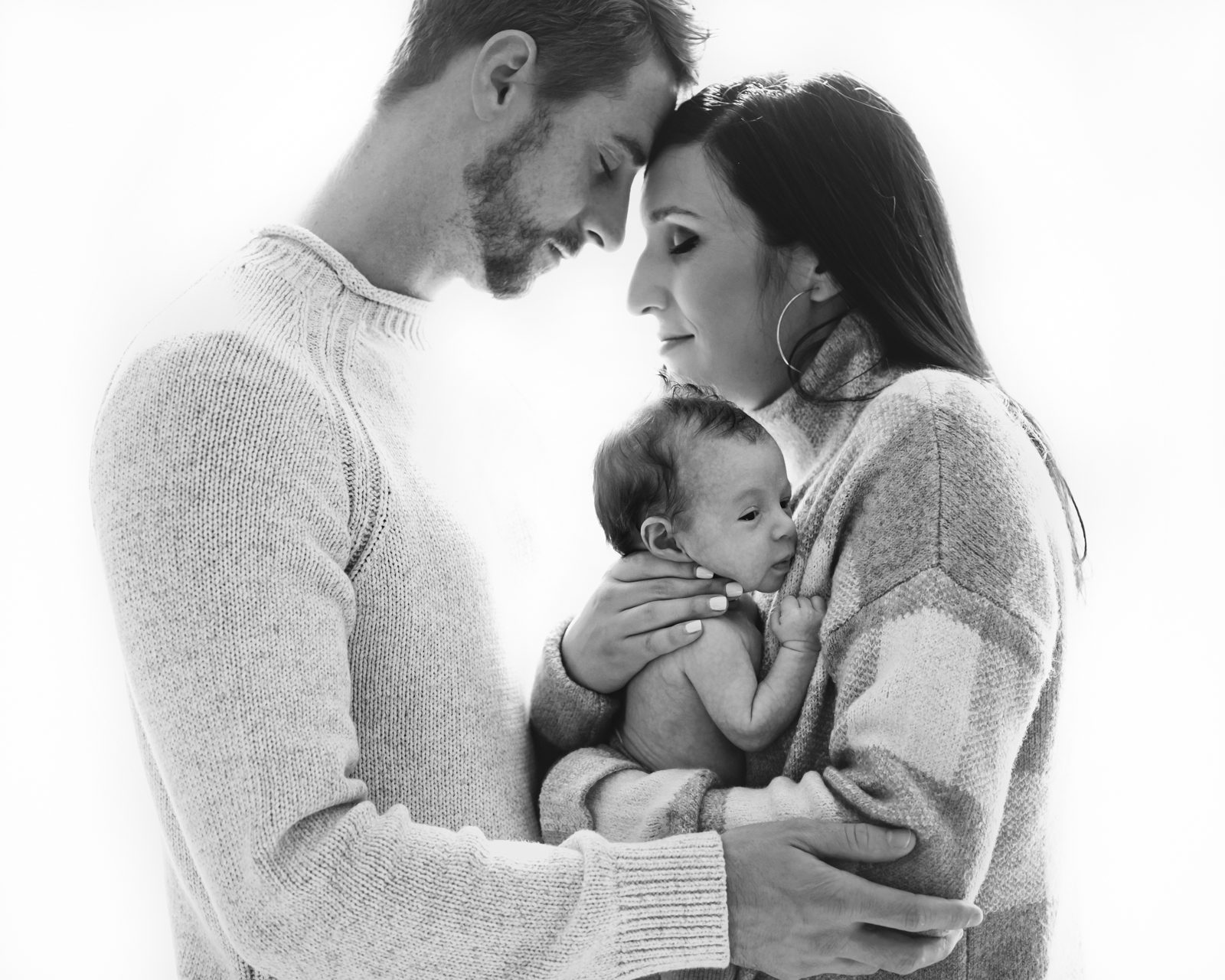 a black and white backlit photo of parents holding their baby boy in between them and resting their foreheads together during a natural light newborn photoshoot