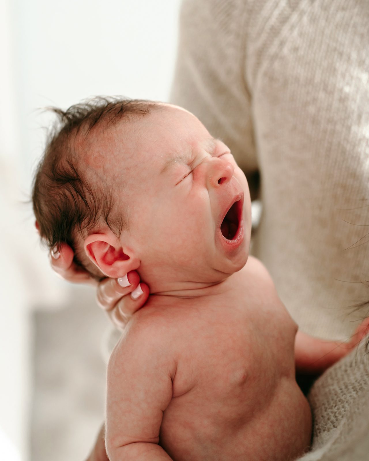 a close up picture of a baby boy yawning while his parents hold him in their arms during a natural light newborn photoshoot