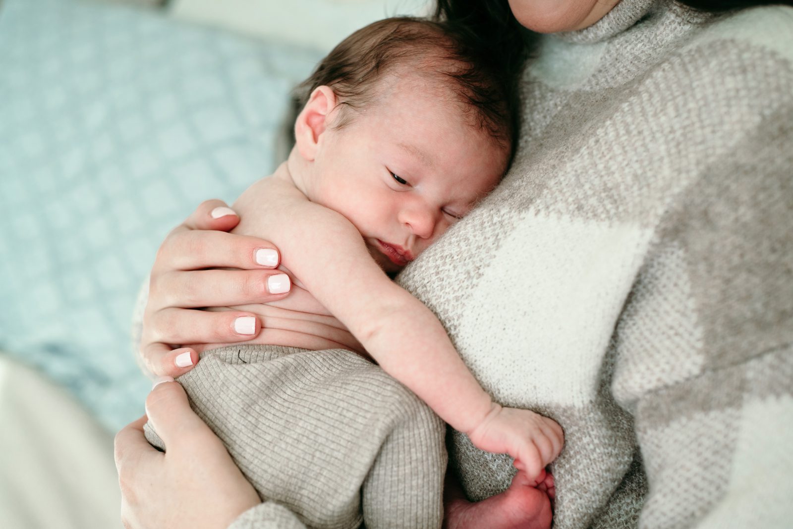 a baby boy snuggled up against his mother's chest and grabbing his toes with his hand during an in home lifestyle newborn photoshoot