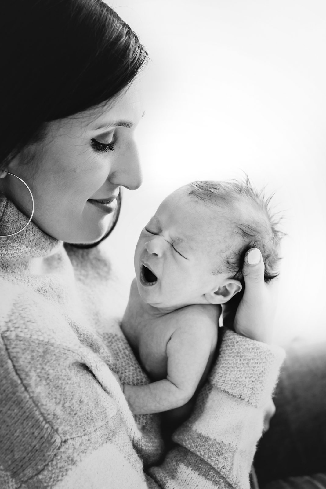 a black and white photo of a mom holding her newborn son against her chest and smiling down at him as he yawns during a natural light newborn photoshoot