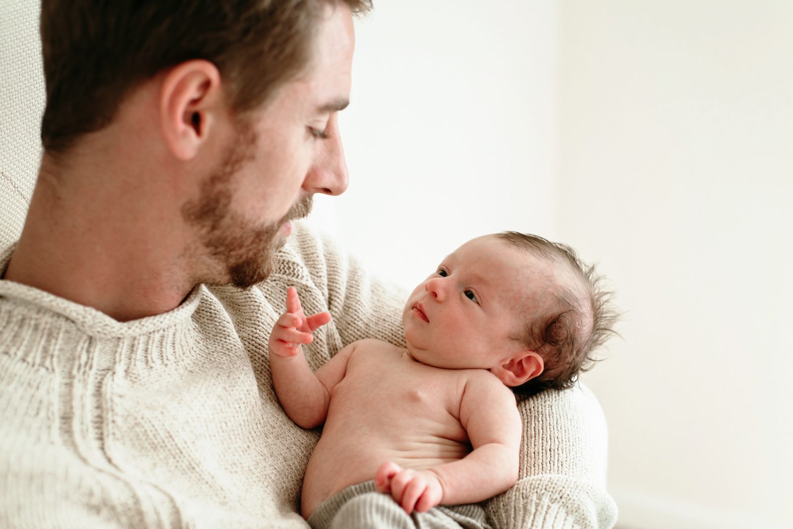 a dad holding his baby boy in his arms as his son looks up at him and reaches toward dad's face with his hand during a natural newborn photoshoot