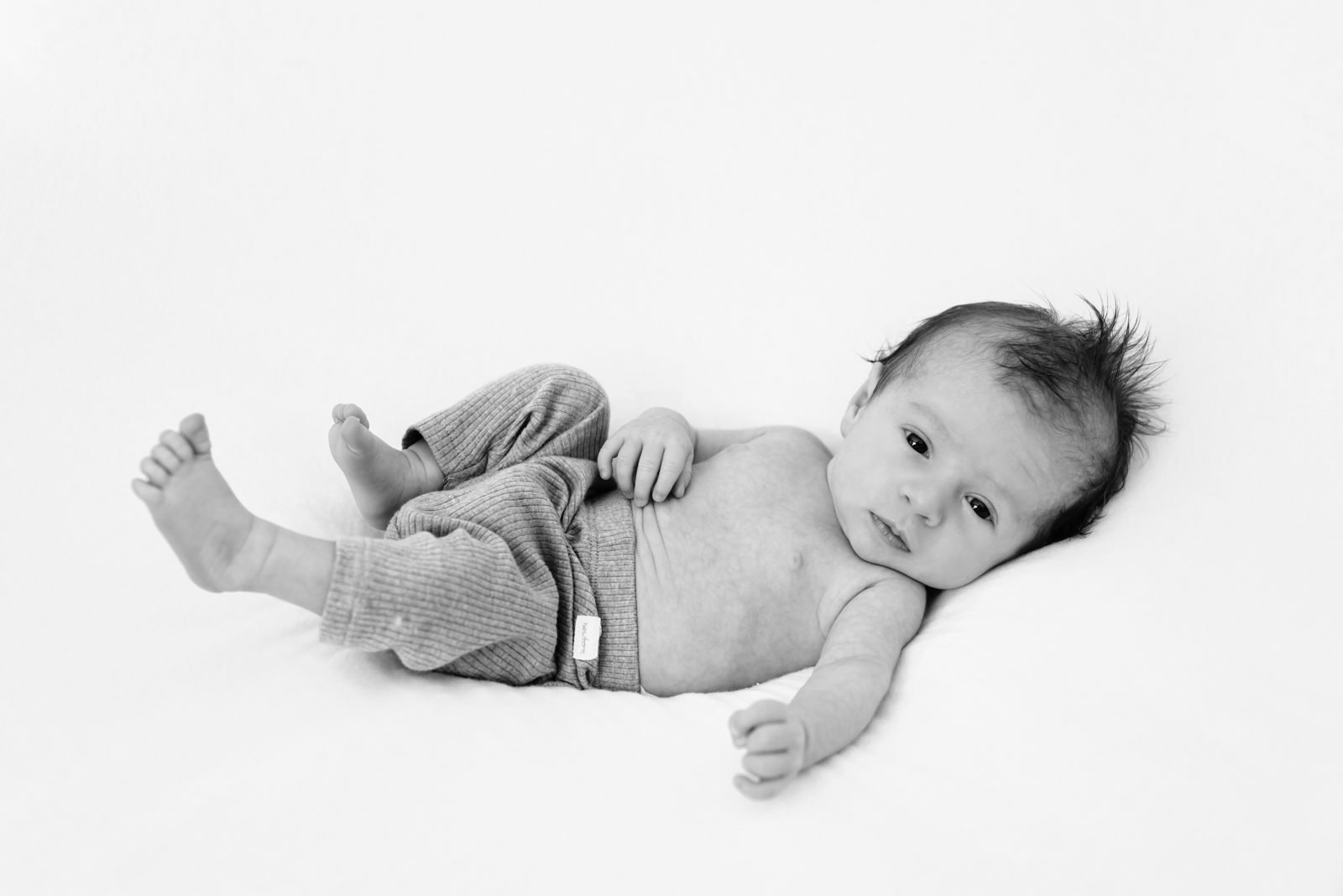 a black and white picture of a baby boy laying on a white backdrop and looking at the camera during a newborn photoshoot