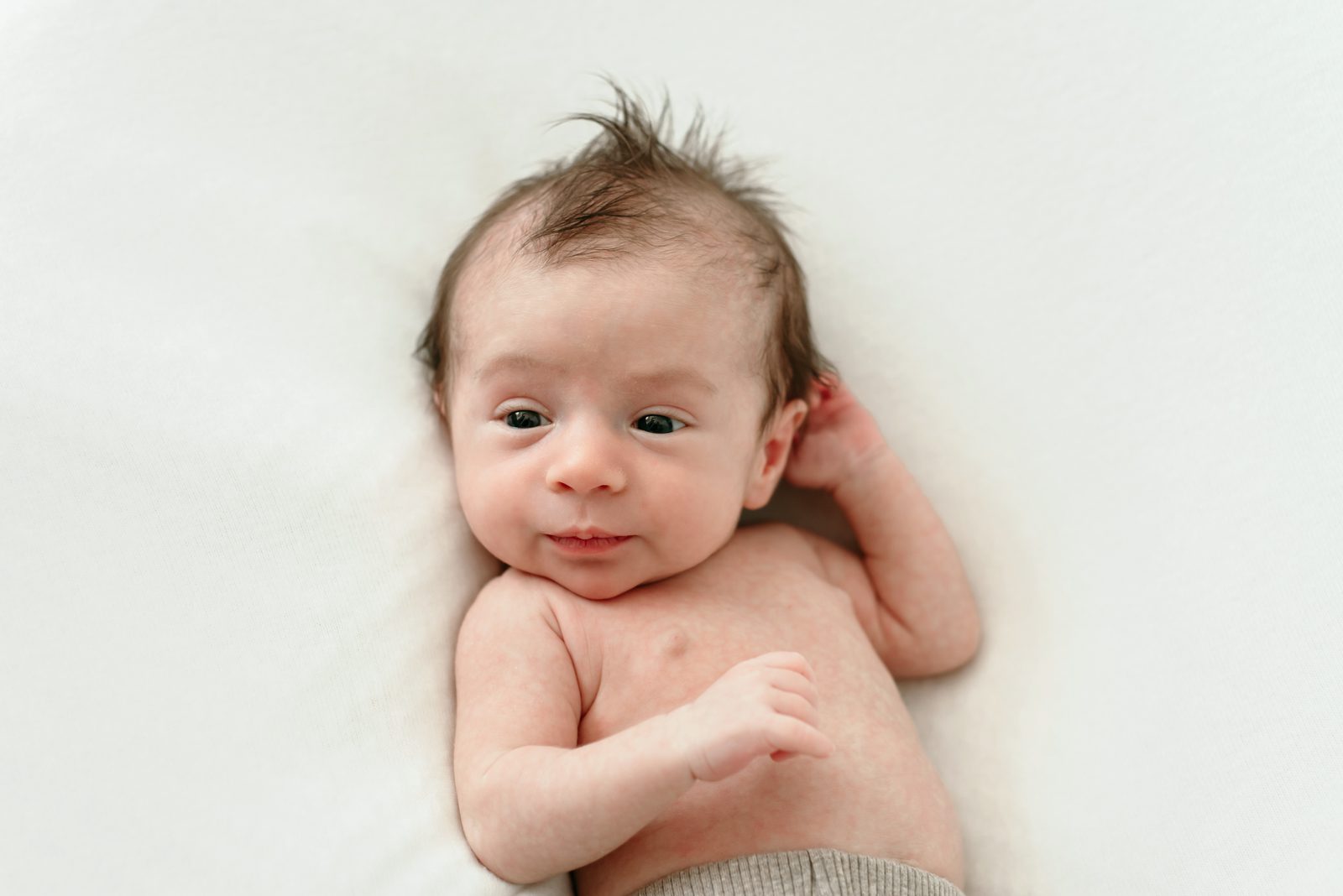 a baby boy laying on a white backdrop and looking off to the side toward the window light during a natural light newborn photo session