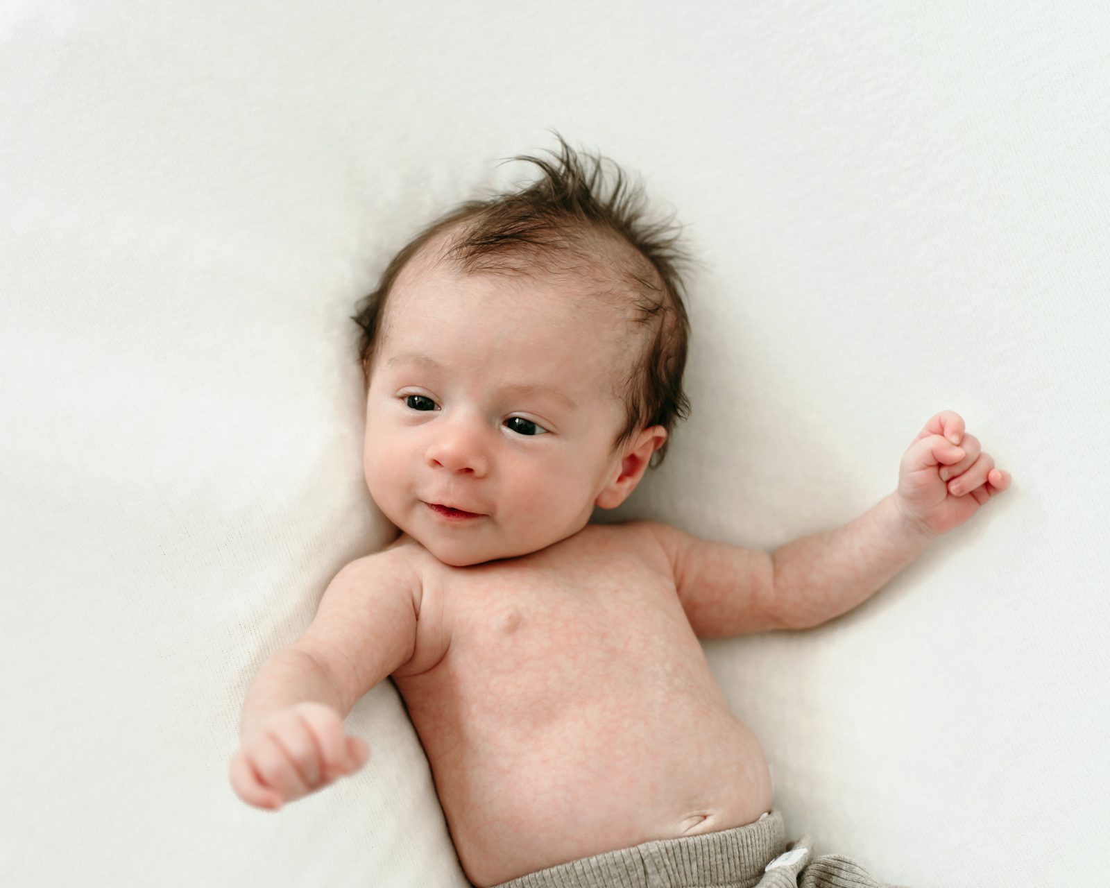 a baby boy laying on a white backdrop and moving his arms around as he looks off to the side and smiles at his parents during a newborn photo session