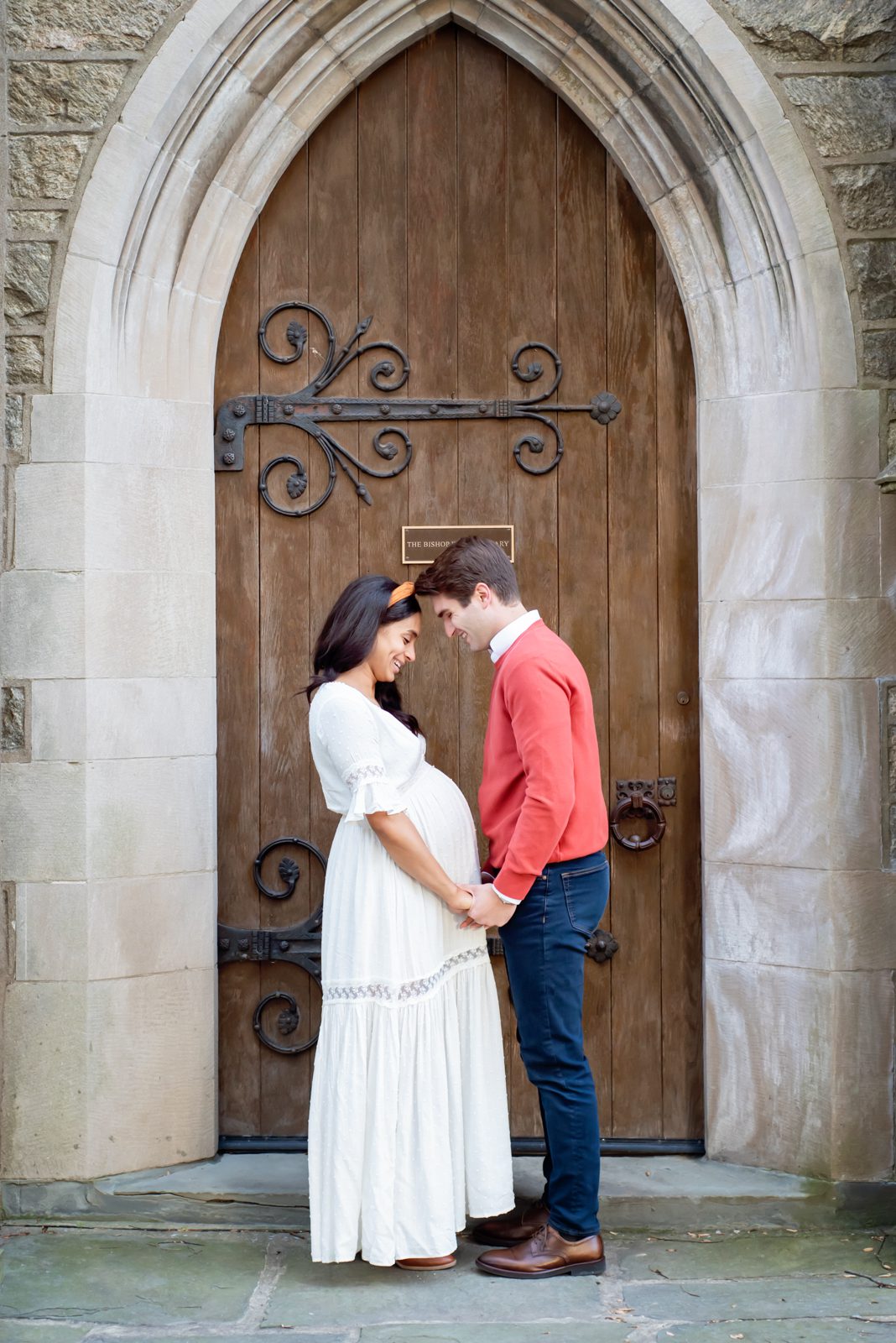 an expecting couple standing in a stone archway and holding hands while they smile down at mom's belly during a maternity photoshoot