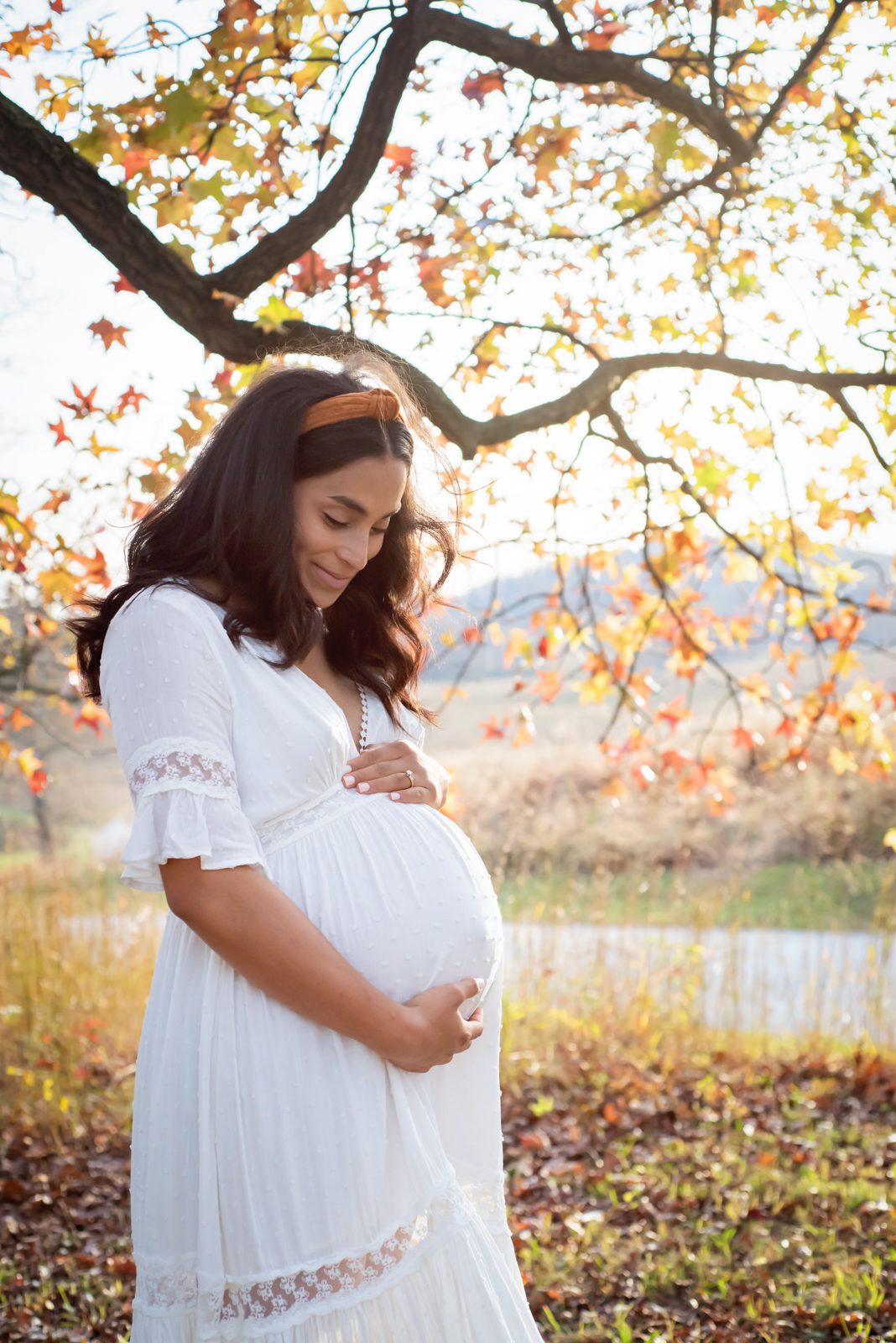 a pregnant mom standing in front of a tree full of orange and yellow leaves and smiling down at her belly during a maternity photoshoot