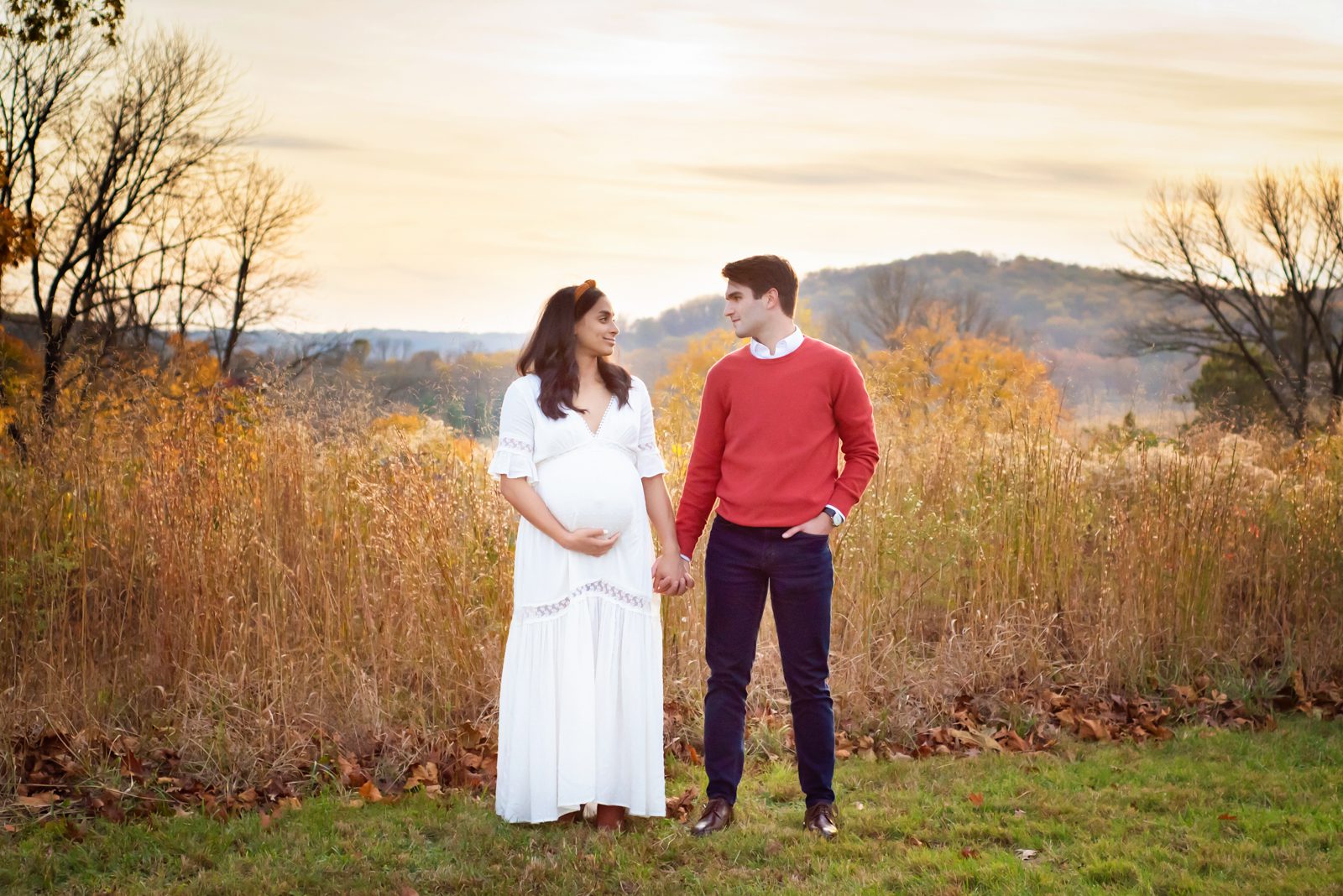 an expecting couple standing in front of tall golden grasses holding hands and smiling at each other during a maternity photo session