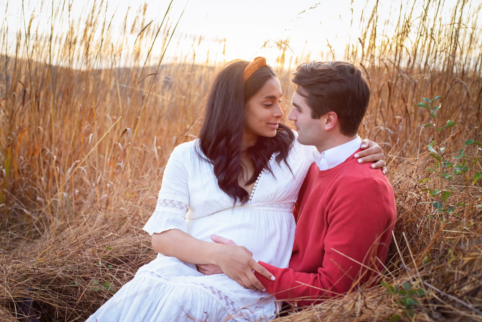 a pregnant mom sitting on her husband's lap in a field of golden grasses and smiling at him during a Valley Forge maternity photo session