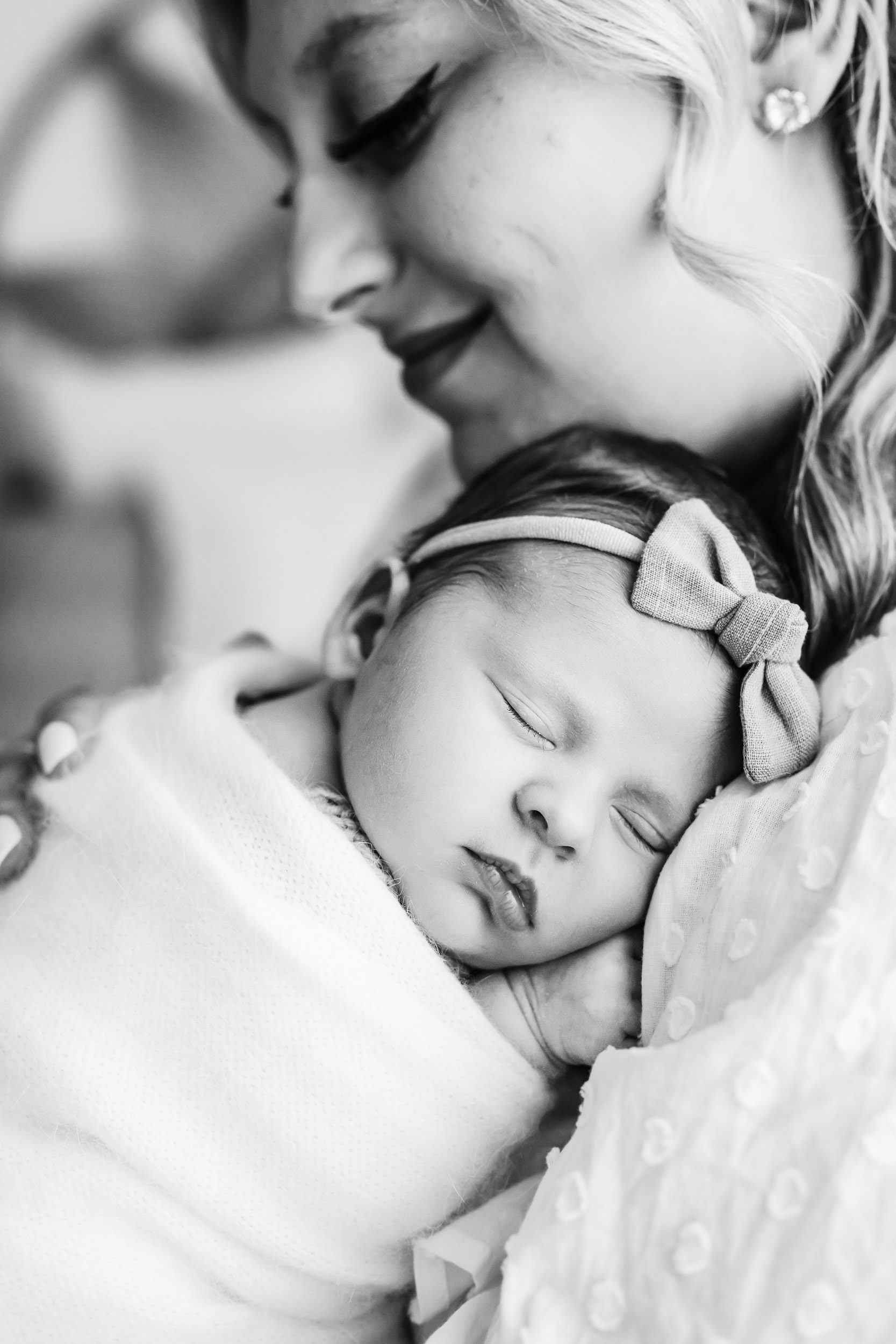 a black and white picture of a baby girl wrapped in a white swaddle blanket sleeping peacefully cuddled up against her mom's chest during a natural newborn photography session