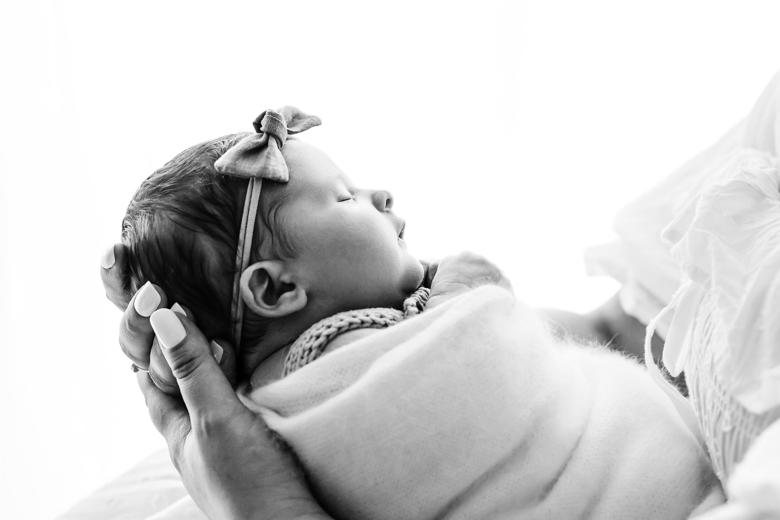 a close up black and white backlit photo of a baby girl swaddled up and sleeping peacefully in her mom's arms during a natural newborn photography session